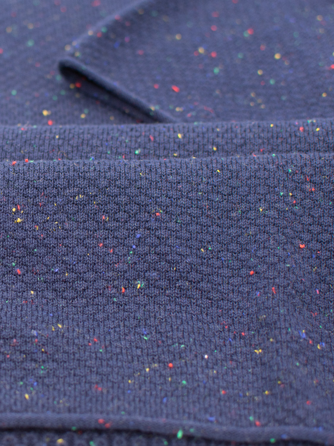 Close-up of the Denver Speckled Knit Cotton Polo by ID Menswear reveals a folded, textured dark blue cotton-knit fabric with small colorful speckles in red, yellow, and green, perfect for casual wear.