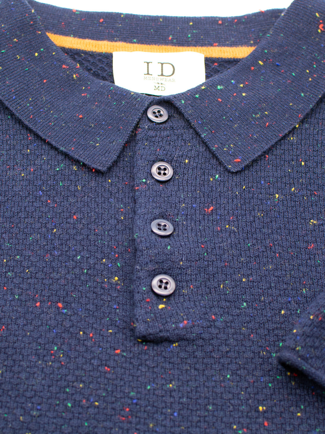 Close-up of the Denver Speckled Knit Cotton Polo by ID Menswear, featuring a navy blue textured cotton-knit with multicolored speckles. It has a collar, five dark buttons and a fabric label showing ID and Medium size for a breathable fit perfect for casual wear.
