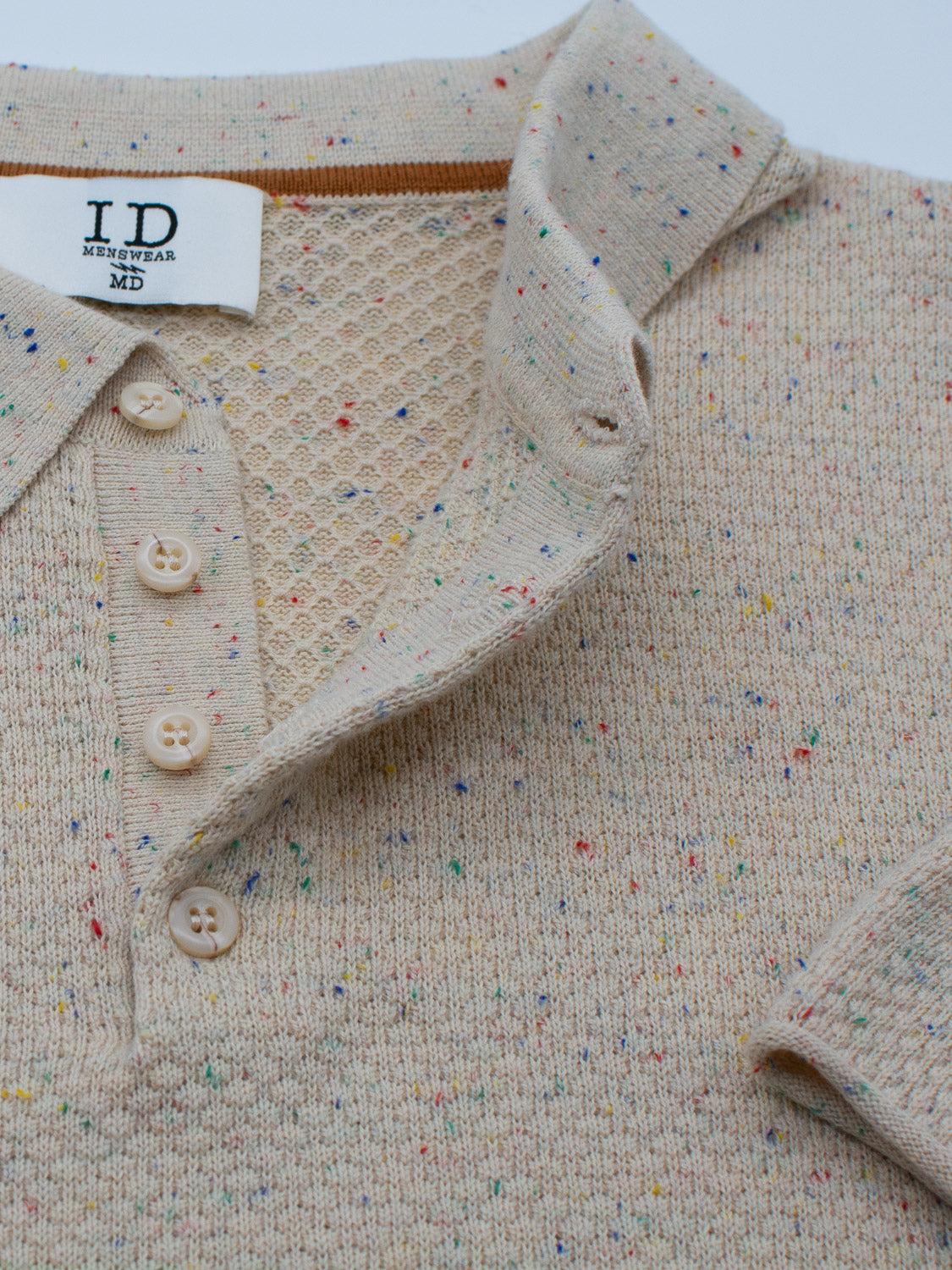 The Denver Speckled Knit Cotton Polo by ID Menswear is a beige, speckled cotton-knit shirt with a honeycomb-patterned interior collar. It has three cream buttons, a fabric ID Menswear tag marked MD, and multicolored specks for a soft texture and breathable casual fit.