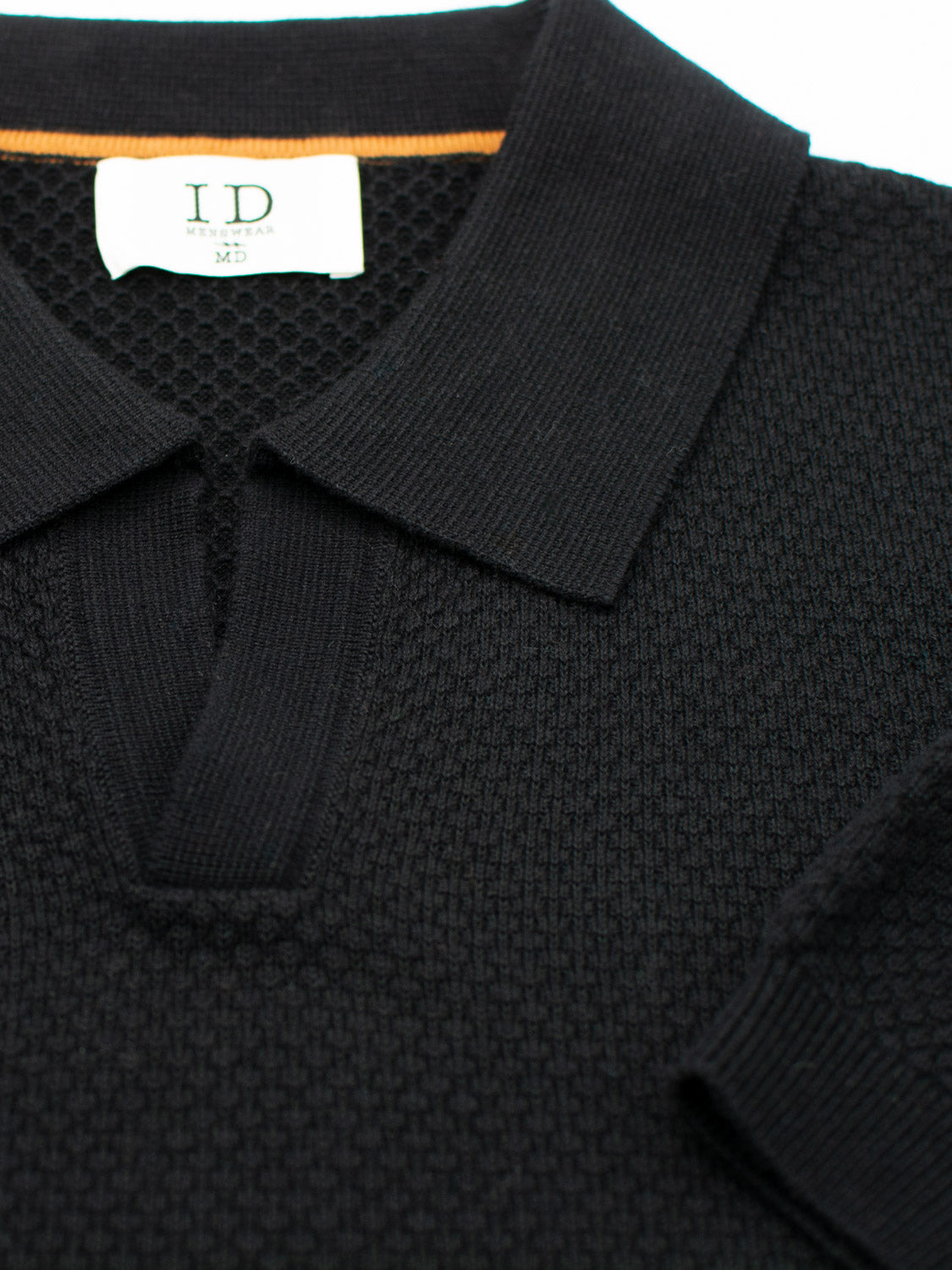 A close-up of the black Malaga Knitted Cotton Polo by ID Menswear, featuring a collared neckline and timeless elegance. The label confirms 100% cotton and size MD. A small orange stripe peeks from the collar as the shirt is displayed flat on a white surface.