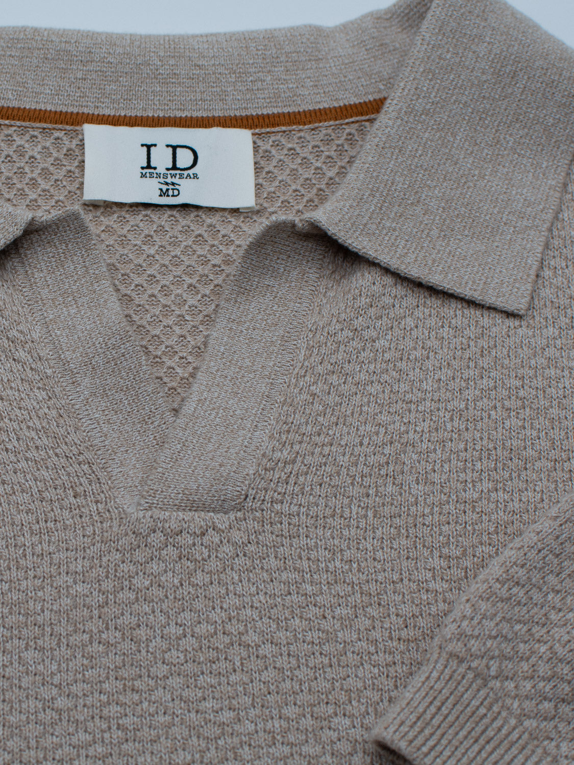 Close-up of the Malaga Knitted Cotton Polo, a beige V-neck sweater with a collar from ID Menswear. Made of 100% cotton with a textured design, it exudes timeless elegance and features a clothing label reading I.D. Menswear MD.