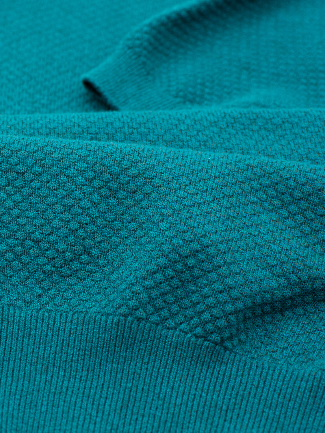 A close-up of the Malaga Knitted Cotton Polo by ID Menswear reveals 100% cotton teal fabric with textured weaving. The polo features distinct diagonal lines and a subtle, raised texture, epitomizing timeless elegance and sophistication.