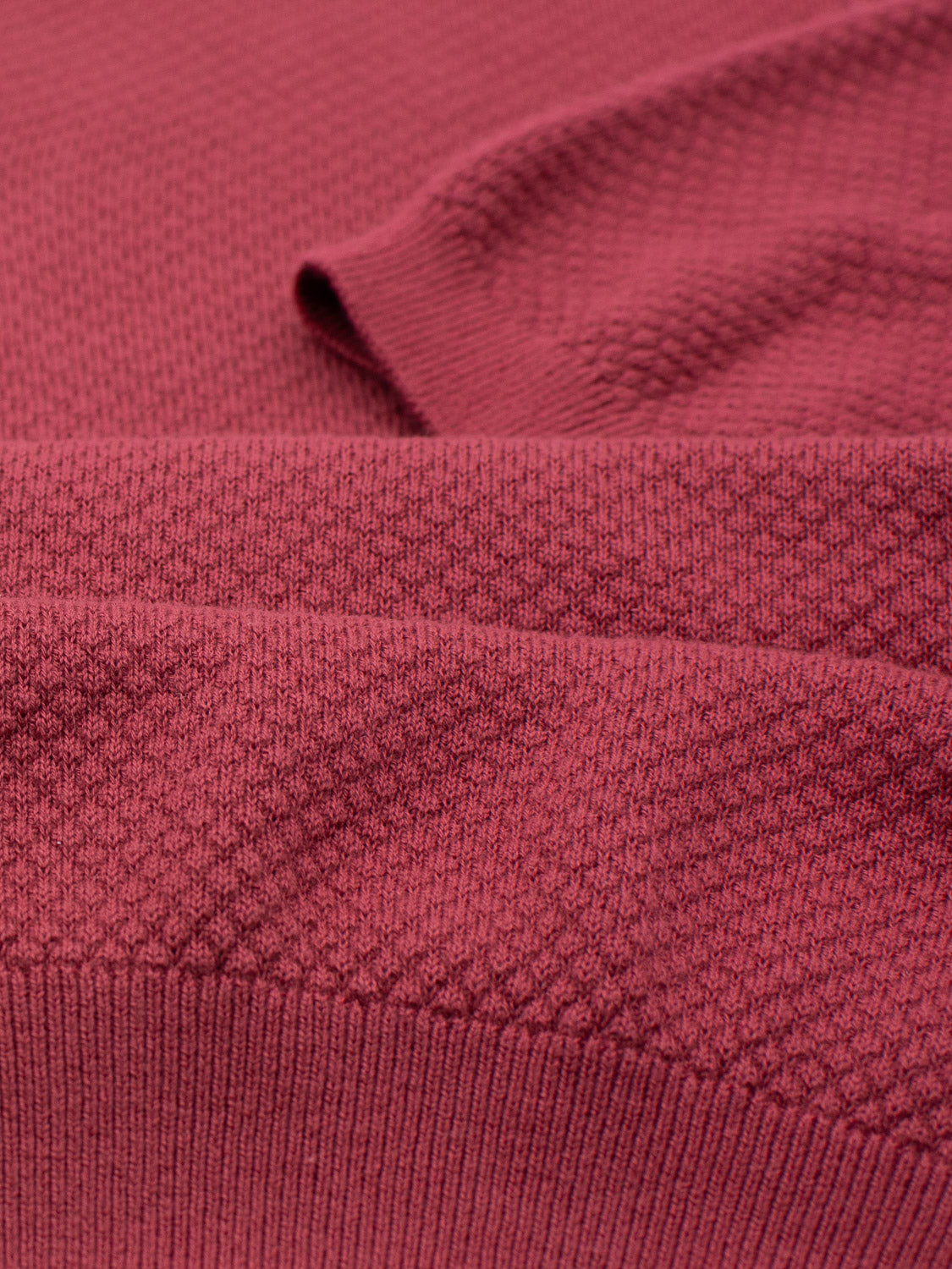 A close-up of the folded red textured fabric exudes timeless elegance with a subtle diamond pattern. The Malaga Knitted Cotton Polo, by ID Menswear, creates soft shadows on its woven details and rich color. Made from 100% cotton, it promises luxury and comfort.