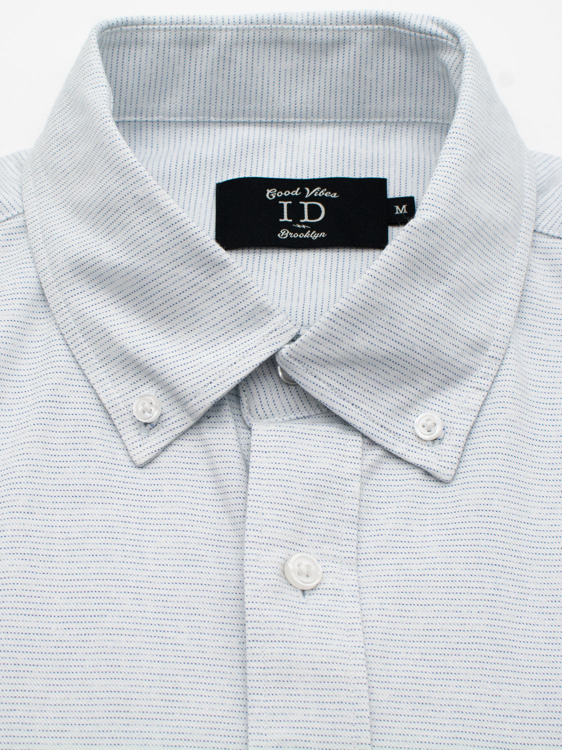 Close-up of the Akira Cotton Button Down Shirt by ID Menswear, a light gray textured cotton shirt with a button-down collar. This regular-fit design features small white buttons and a black label inside the collar, reading Cool White ID Brooklyn in size M.