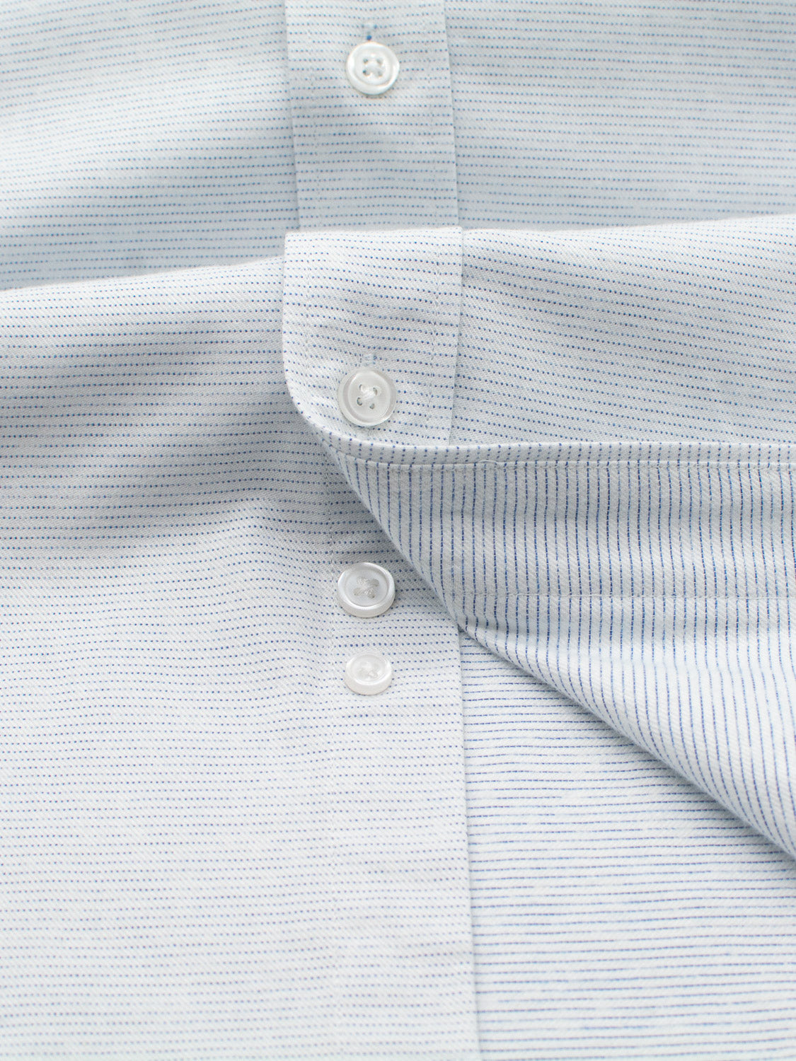 Close-up of an Akira Cotton Button Down Shirt by ID Menswear, featuring a stylish light blue and white striped cotton with a buttoned flap. The fabric has thin vertical stripes and three visible white buttons, slightly folded at the bottom right corner for a regular fit.