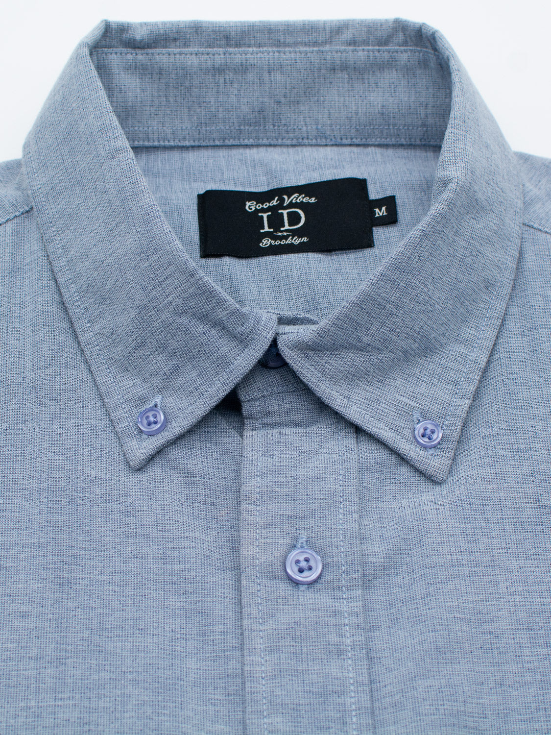 Close-up of a folded light blue Akira Cotton Button Down Shirt made from textured cotton. Featuring a button-down collar, it is labeled ID Menswear in black alongside Good Vibes Brooklyn and size M, all presented against a white background.
