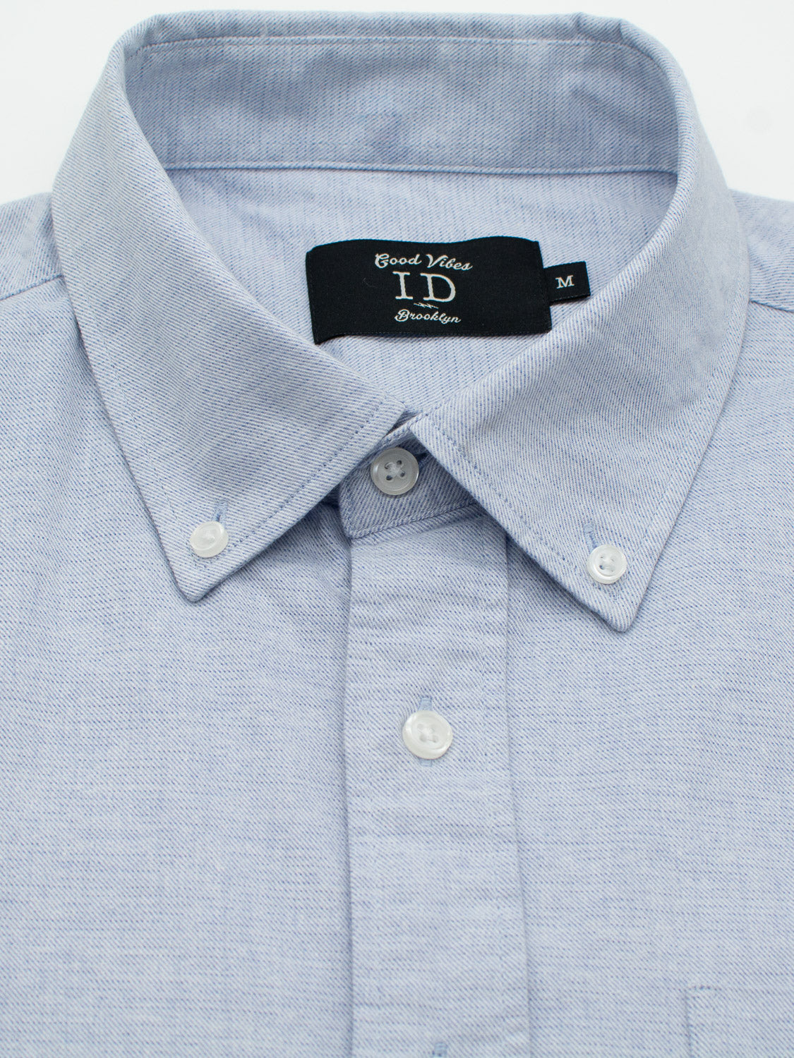 Close-up of a light blue, regular-fit Akira Cotton Button Down Shirt by ID Menswear, featuring a soft textured fabric and white buttons on a button-down collar. The black label reads Good Vibes, ID, Brooklyn in white lettering.