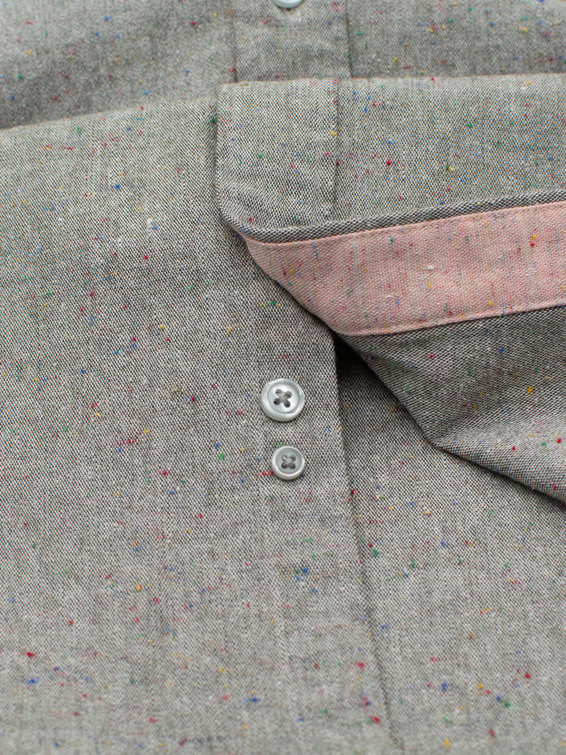 A close-up of ID Menswears Allswell Speckled Button Down Shirt highlights its versatile design with gray fabric, multicolored speckles, and two white buttons. A touch of pink lining adds flair, showcasing the premium cotton-linen blend.
