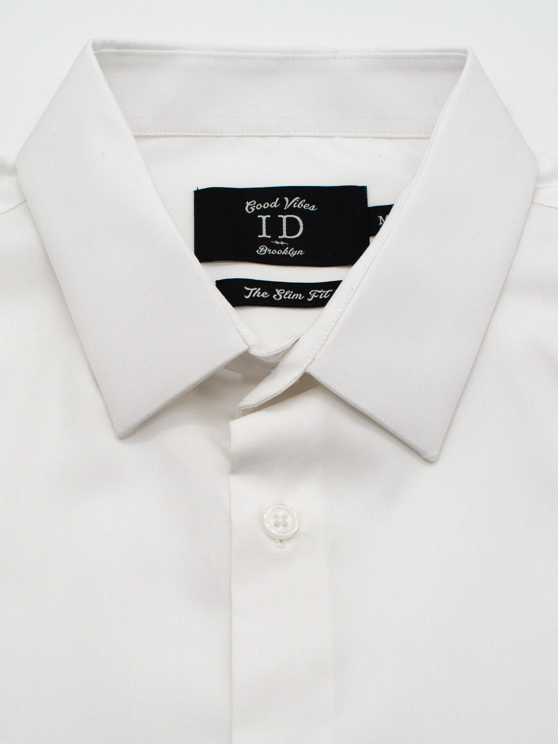 Close-up of a white, long-sleeve dress shirt with a pointed collar. It features a button-up front and inside, a black tag displays ID Menswear along with Good Vibes and The Slim Fit in white text. Product: ID Cotton Dress Shirt in Slim and Regular Fit.