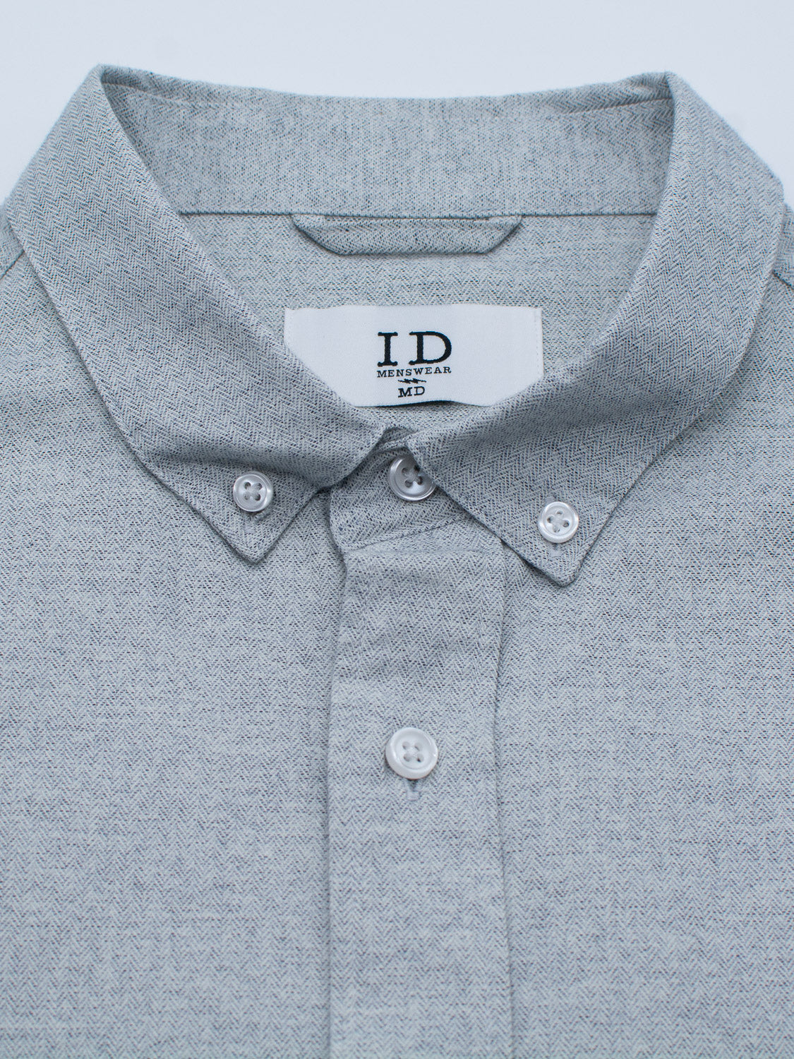 Close-up of the Maya Fine Herringbone Shirt by ID Menswear, a light gray button-down crafted from 100% cotton with a subtle herringbone pattern. It features white buttons, a button-down collar, an I D Menswear label inside the collar, and soft textured fabric.