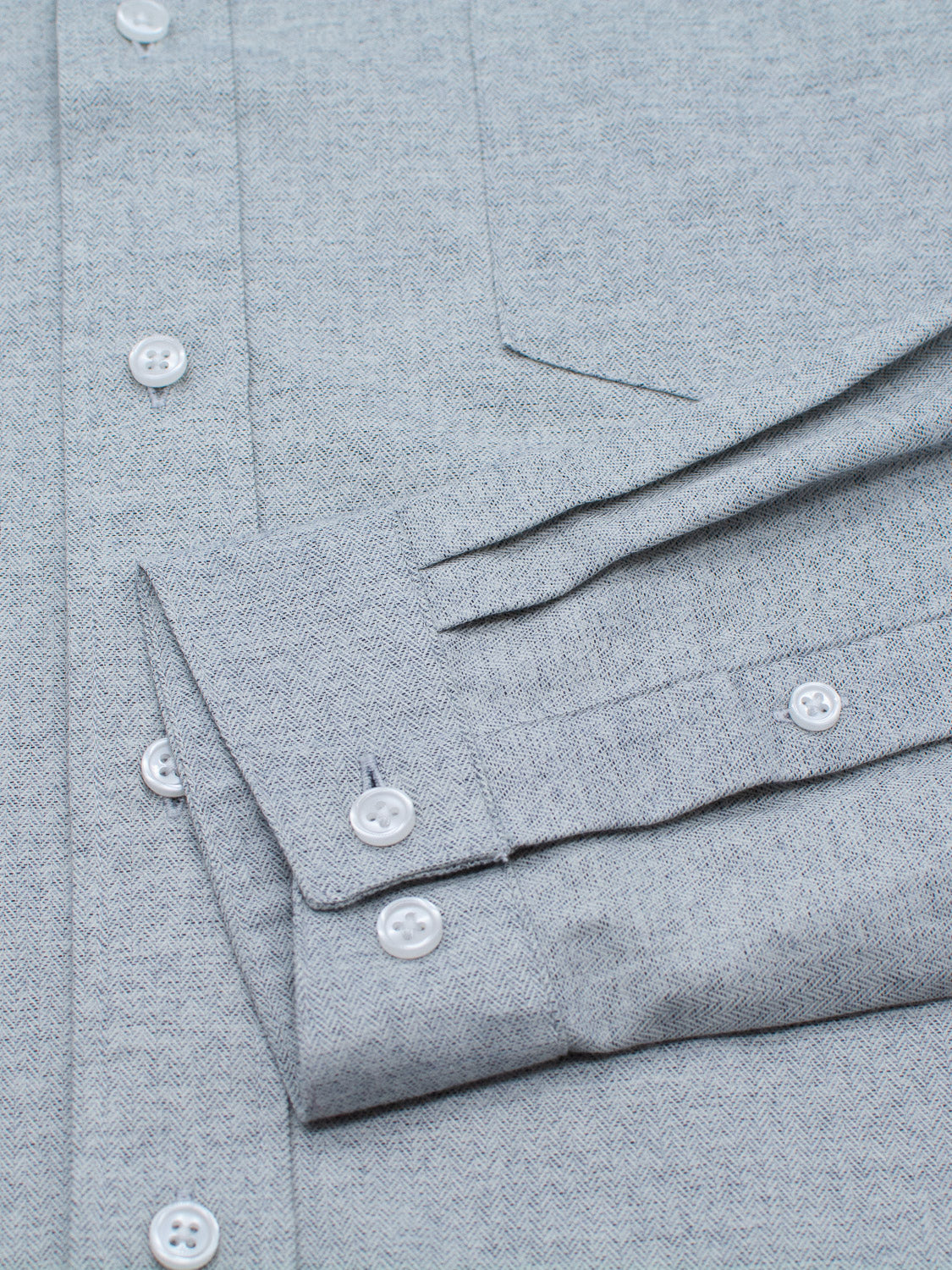 The Maya Fine Herringbone Shirt by ID Menswear features a gray flannel design with white buttons and a folded sleeve showcasing a buttoned cuff. The 100% cotton fabric and button details enhance this stylish button-down shirt.