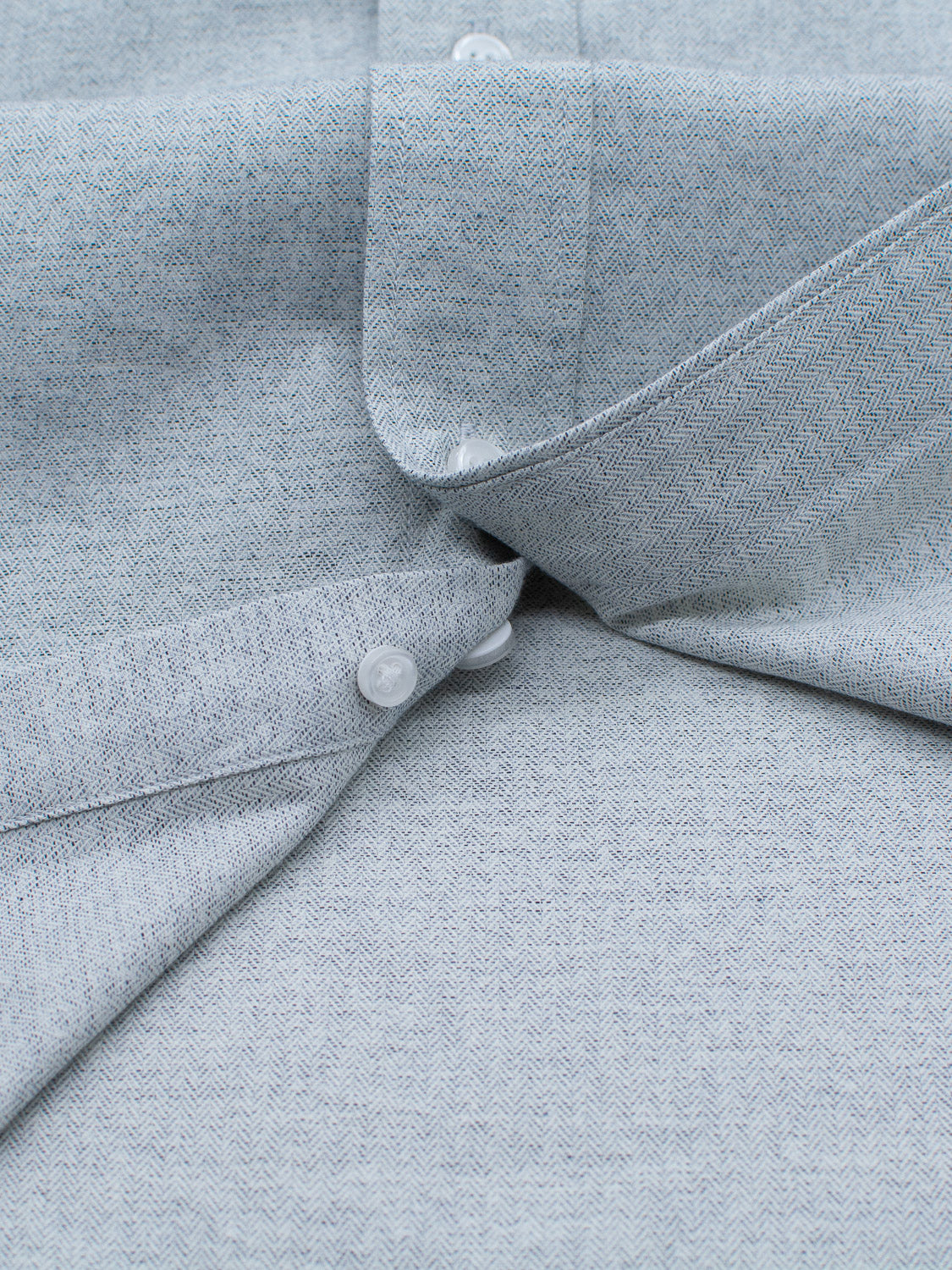 Close-up of the 100% cotton Maya Fine Herringbone Shirt by ID Menswear in light blue-gray, featuring white buttons. One button is fastened while another is unfastened, highlighting the shirts subtle herringbone texture.