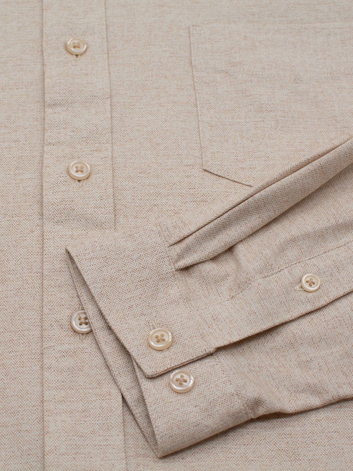 Close-up of the Maya Fine Herringbone Shirt by ID Menswear, showcasing its 100% beige brushed cotton fabric with pocket and button details. The neatly folded sleeves feature matching buttons, enhancing the classic design with its textured appearance.