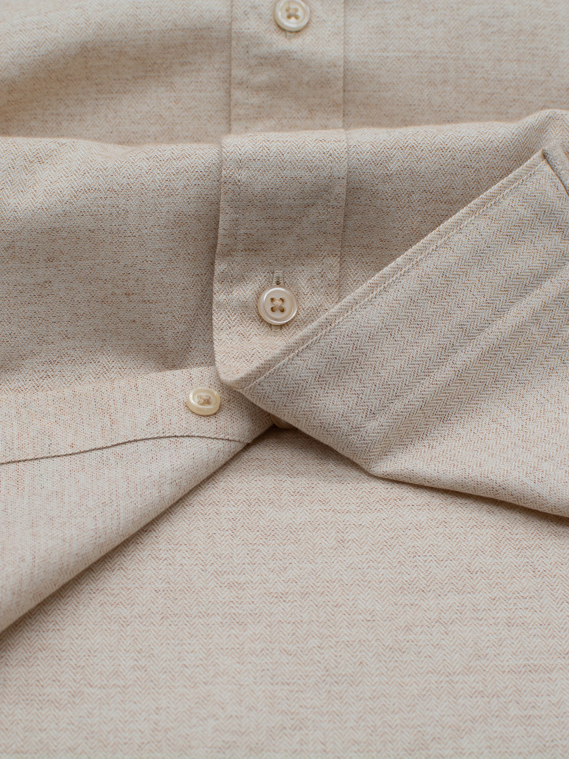 A close-up of the Maya Fine Herringbone Shirt by ID Menswear shows beige fabric with a herringbone pattern, highlighting the sleeve and buttons. The texture and stitching are clear, emphasizing its 100% cotton composition.