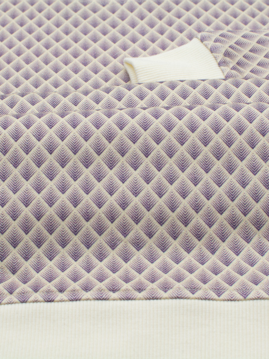 A close-up of the Carp Geometric Printed Sweatshirt from ID Menswear shows a textured, purple and off-white diamond pattern. The visible edges highlight its construction, crafted from bamboo and organic cotton fleece, giving it a modern artistic vibe.