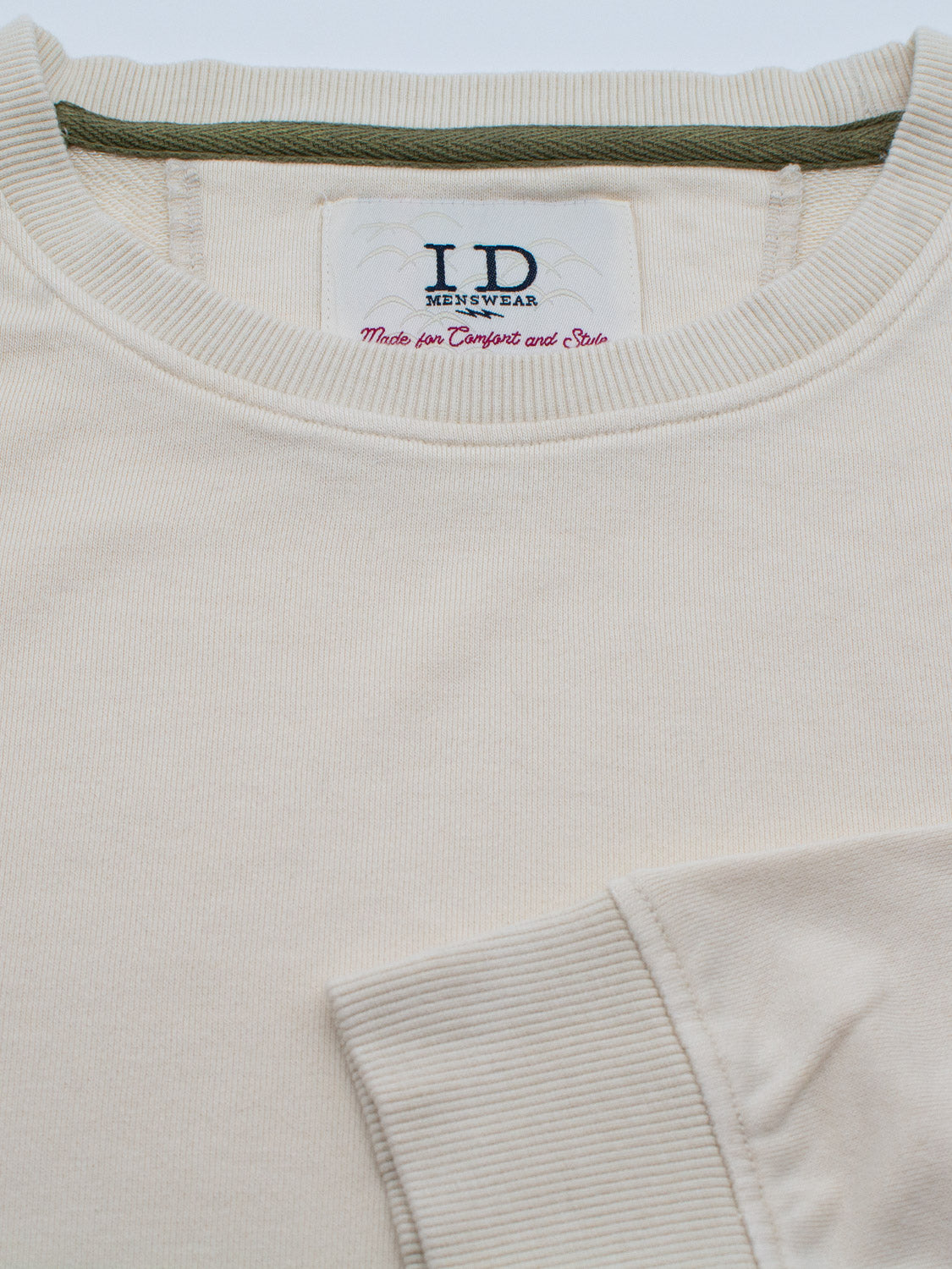 Close-up of the Champ Bamboo Organic Cotton Sweatshirt by ID Menswear, featuring a beige crewneck with green trim on the collar. A visible tag reads Made for Comfort and Style, while a sleeve in the foreground highlights its soft texture.