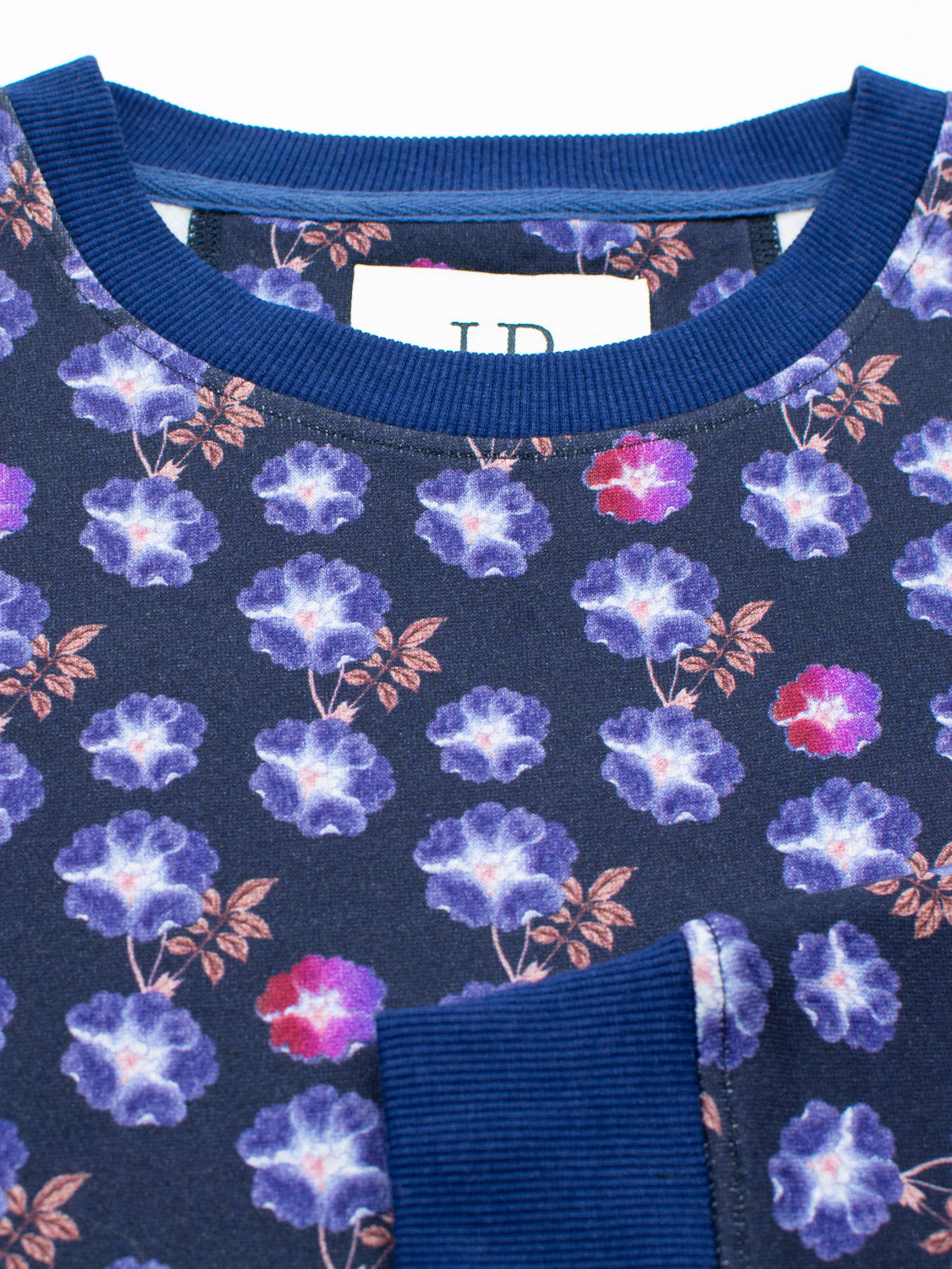 Close-up of the Flora Printed Sweatshirt by ID Menswear, featuring a dark blue fabric with vibrant purple and pink floral digital print and brown leaves. The pattern adorns a garment with a visible seam, small pocket, and partial label.