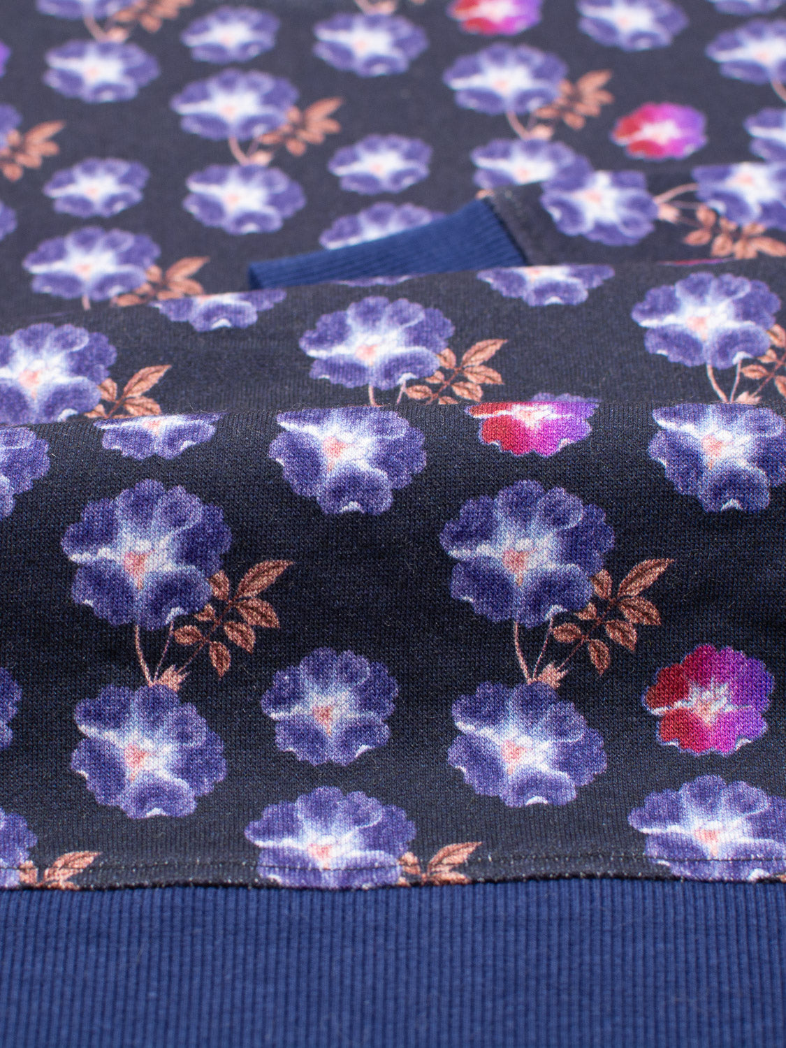 A close-up of ID Menswears Flora Printed Sweatshirt reveals a vibrant digital design featuring blue, purple, and red flowers with brown leaves on a dark background. A blue ribbed section at the bottom indicates it’s crafted from bamboo and organic cotton fleece.