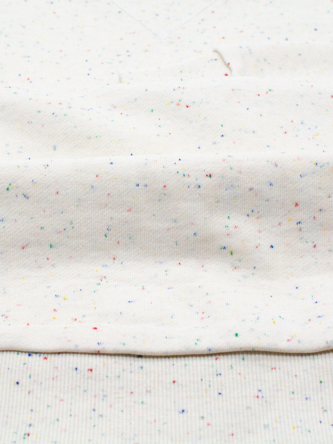 Close-up of the Stardust Speckled Sweatshirt by ID Menswear, featuring a light beige fabric with multicolored specks and ribbed texture. The folded cotton yarn displays gentle curves and shadows, enhancing the colorful stardust effect throughout.