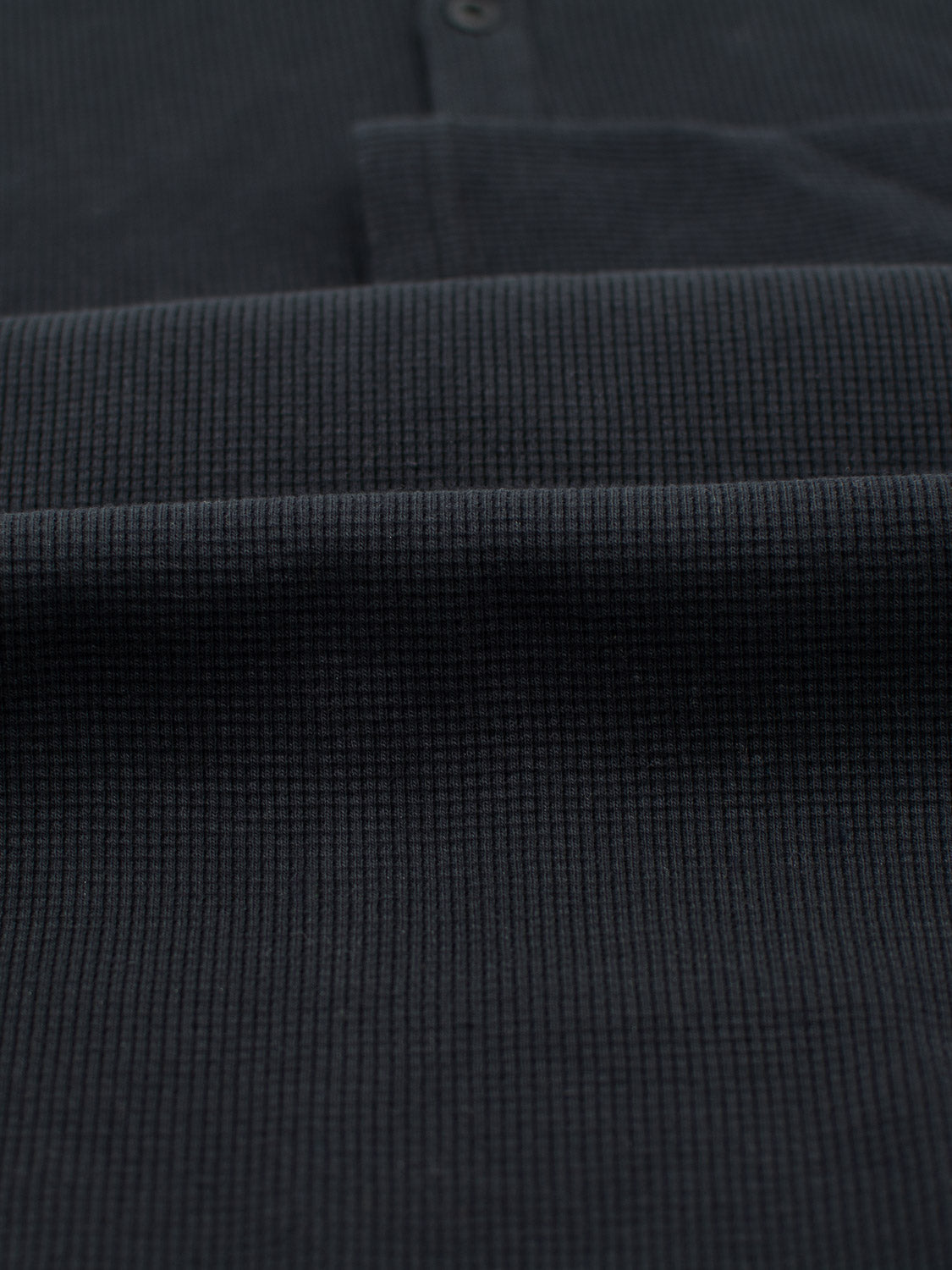 Close-up of ID Menswears Freestyle Waffle Knit Henley, featuring a dark waffle knit with a fine grid pattern. The cotton-spandex blend appears soft, with two buttons partly visible at the top center.
