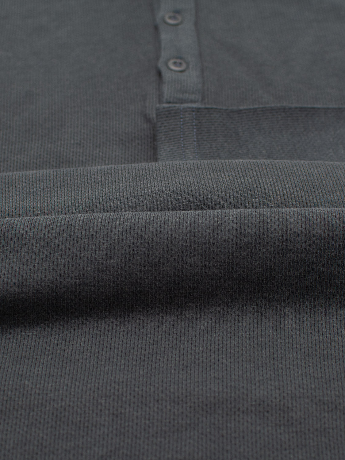Close-up of the Freestyle Honeycomb Knitted Mesh Henley by ID Menswear, showcasing its dark gray ribbed fabric made from organic cotton. It features a three-button placket and horizontal ribbing for subtle charm in this eco-friendly piece.