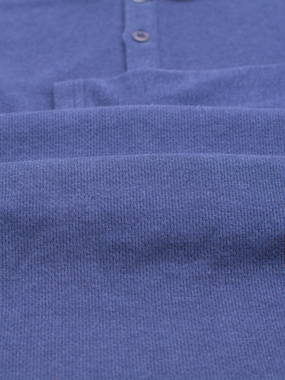 Close-up of folded Freestyle Honeycomb Knitted Mesh Henley by ID Menswear; blue ribbed fabric with buttons suggests a collared garment. The texture and color are showcased, crafted from organic cotton, with neat folds.