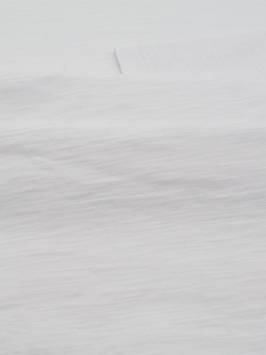 A close-up of the Paradise Long Sleeve Tee by ID Menswear reveals smooth, white slub cotton fabric with subtle horizontal lines. Its lightweight, slightly wrinkled appearance and soft texture make it ideal for casual wear or linens, while gentle lighting enhances its delicate surface.