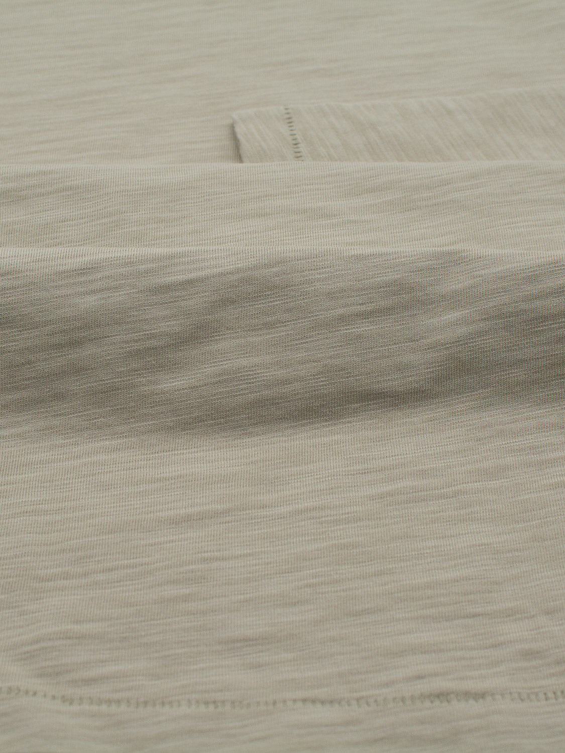 Close-up of folded light gray fabric with a subtle woven texture, ideal for the Paradise Long Sleeve Tee by ID Menswear. Its fine lines and soft sheen hint at the smooth, lightweight feel typical of slub cotton, perfect for crafting casual wear.