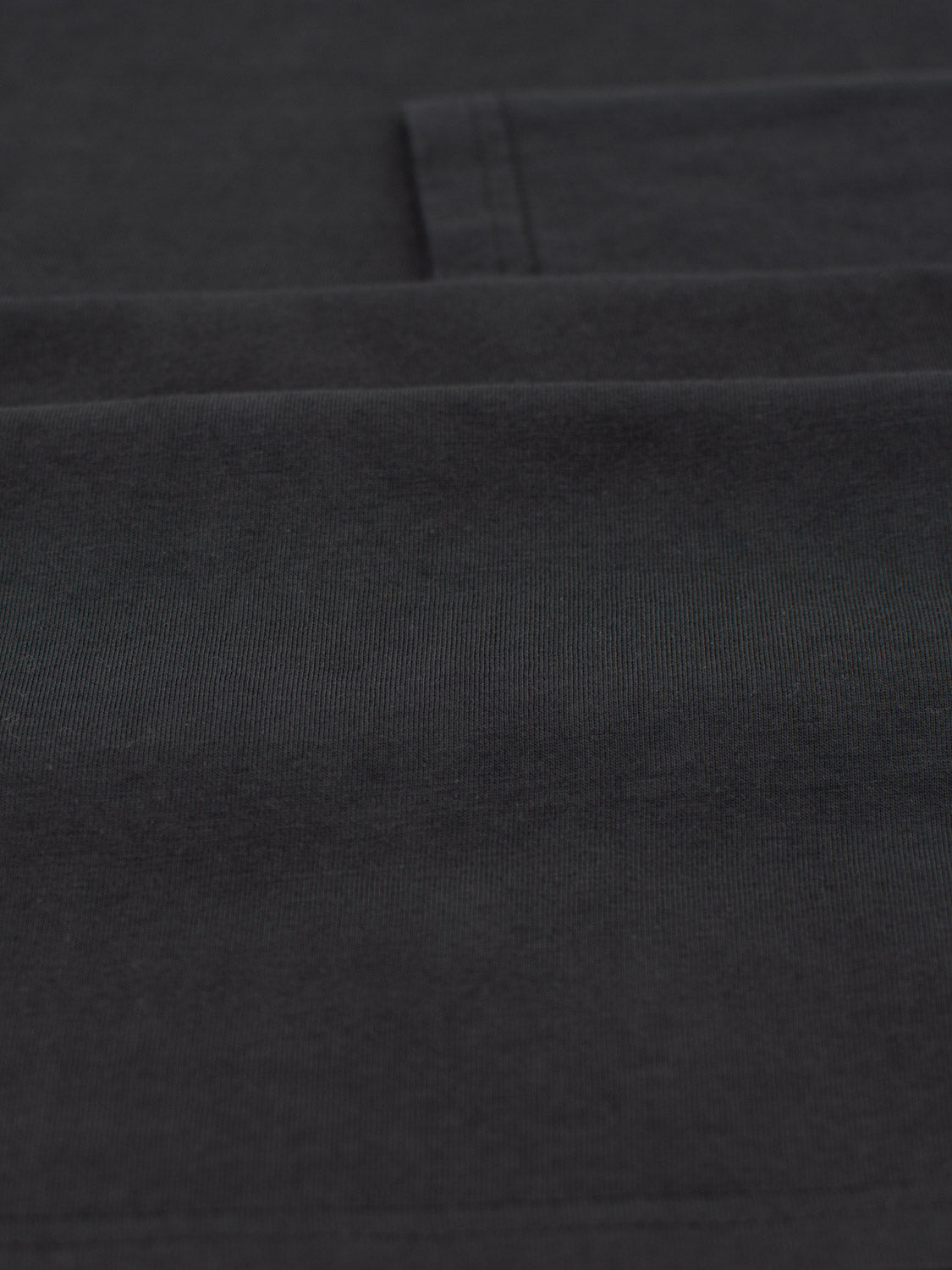 Close-up of a neatly folded Sublime Long Sleeve Tee by ID Menswear, showcasing its sleek dark fabric, visible texture, and seam lines. The smooth, monochrome surface accentuates the fine stitching and craftsmanship, embodying quality casual wear.