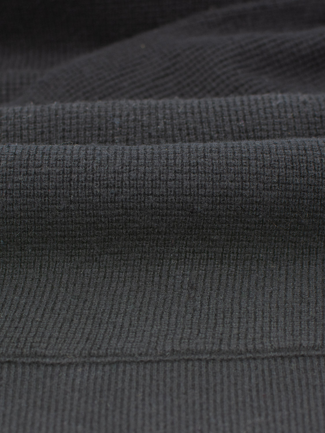 A close-up of the dark, textured fabric reveals visible horizontal and vertical lines, resembling a waffle knit. The material appears soft and slightly folded, highlighting its weave and texture—much like the cozy embrace of ID Menswear’s Bergen Waffle Knit Merino Hoody.