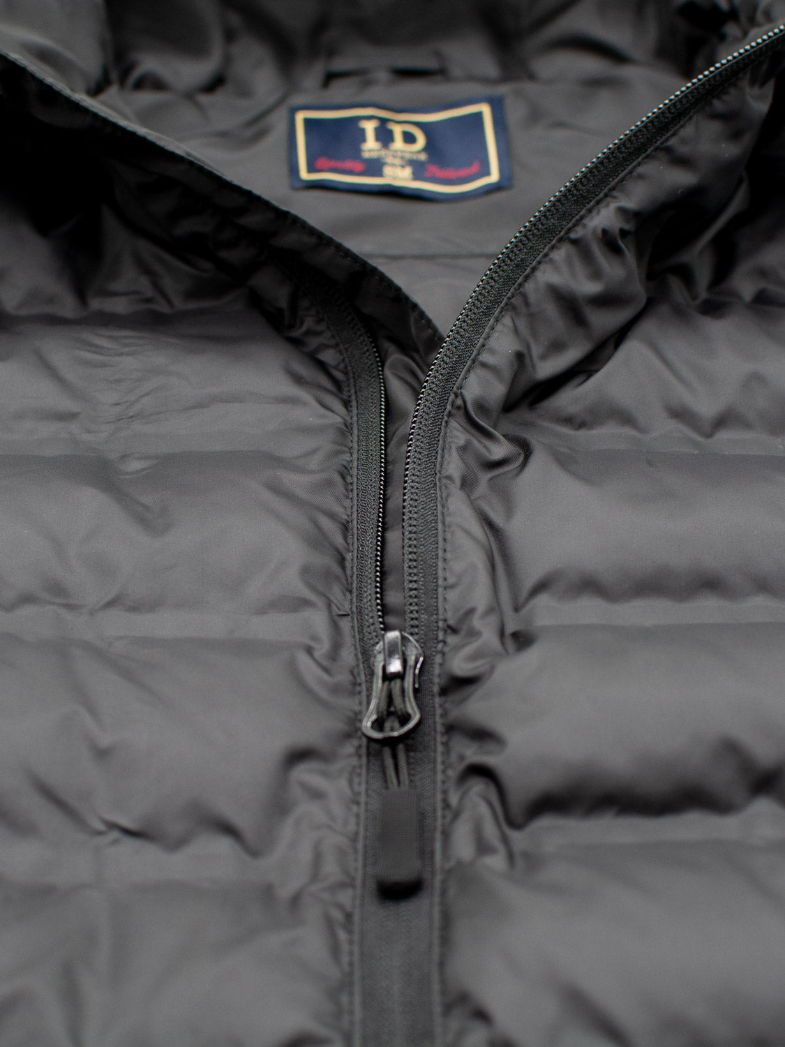 Close-up of the Yukon Hooded Puffer Jacket by ID Menswear, featuring a black lightweight design with a central visible zipper and an inner collar label.