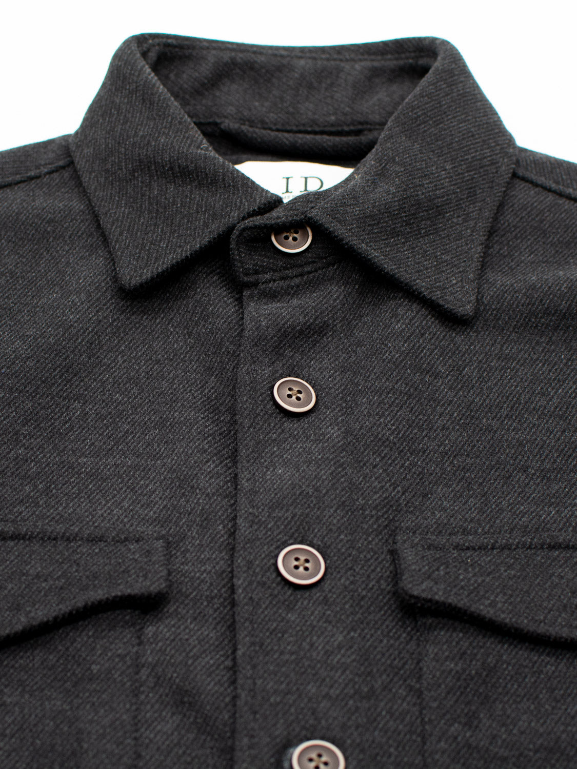 A close-up of the Yukon Shirt Jacket by ID Menswear showcases its dark gray fabric with subtle diagonal texture, ideal for transitional weather. It features brown buttons, two chest pockets, and a partial label with IR inside the collar, making it perfect for casual outings.