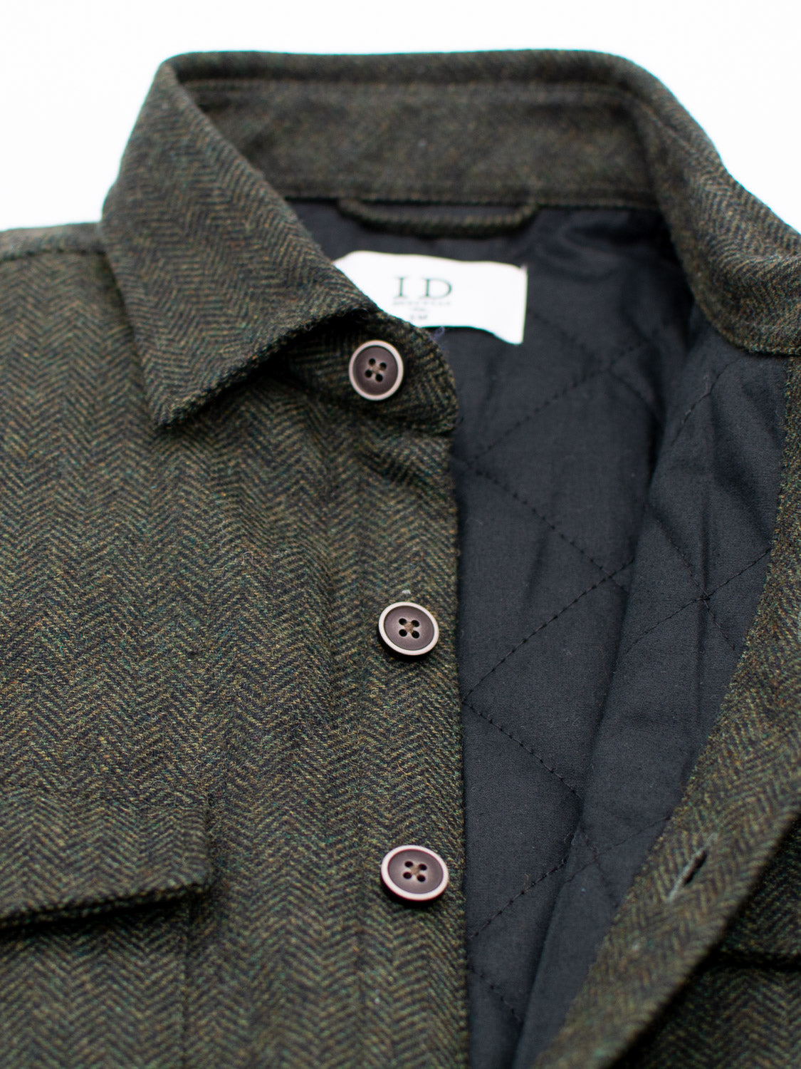 A close-up of the Stuntman Wool Blend Tweed Jacket by ID Menswear, featuring herringbone dark tweed, brown buttons, a pointed collar, black quilted lining, and an “ID.” label that highlights classic outerwear craftsmanship.