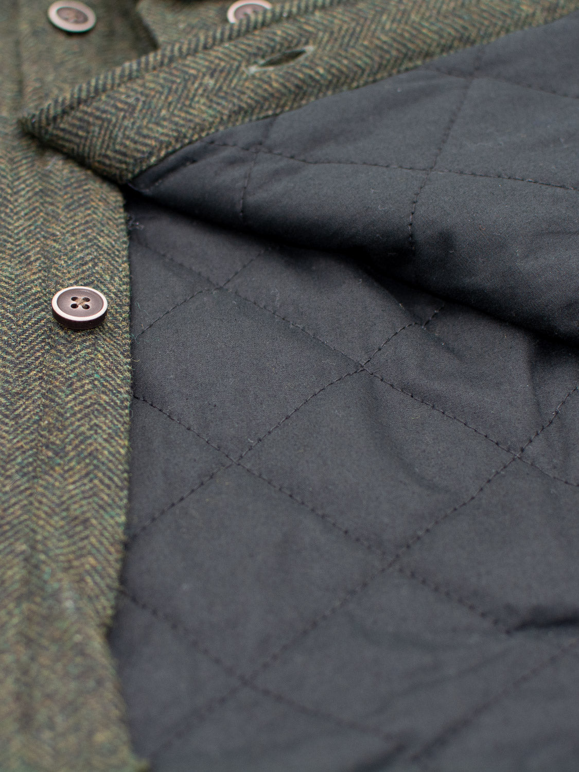 Close-up of the Stuntman Wool Blend Tweed Jacket by ID Menswear, showcasing green herringbone fabric, brown buttons, quilted lining, and visible stitching for a timeless outerwear style.
