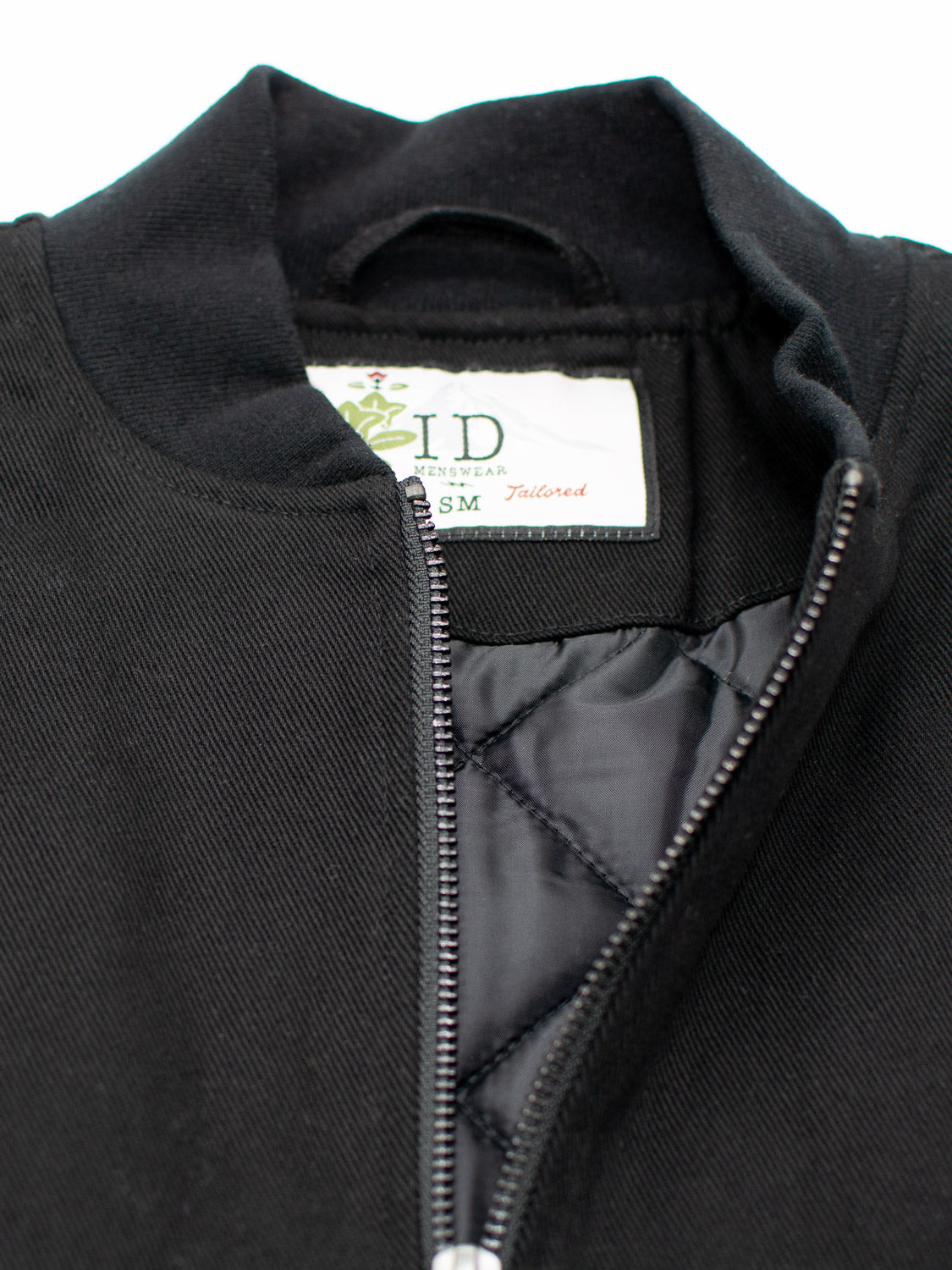 Close-up of Yonkers Cotton Canvas Jacket in black showing a zipper and quilted lining. The ID Menswear label inside indicates Size SM, Tailored, with a small green plant design.