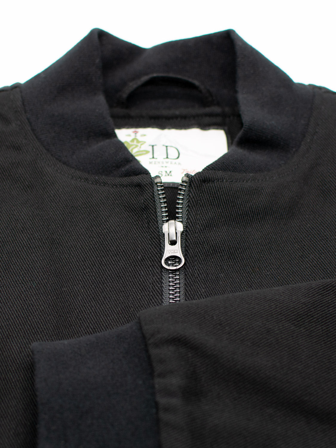 Close-up of the Yonkers Cotton Canvas Jacket by ID Menswear, featuring a central silver zipper partially unzipped to reveal a floral label. The black fabric is ribbed with a round collar and includes cotton-padded quilted lining for warmth.