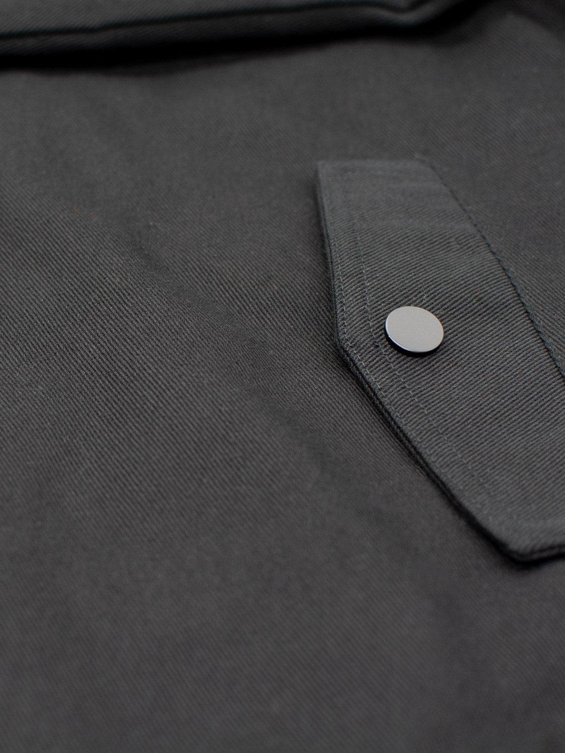 The Yonkers Cotton Canvas Jacket by ID Menswear features a close-up of its dark fabric with a buttoned flap pocket, showcasing intricate stitching and smooth texture. The metallic button adds flair, while the cotton-padded quilted lining ensures warmth and comfort.