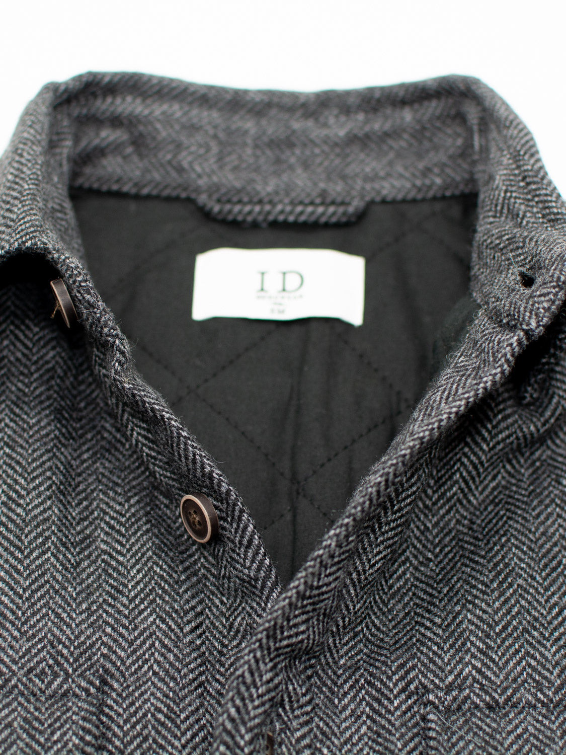 Close-up of the Stuntman Wool Blend Tweed Jacket by ID Menswear, featuring a gray herringbone pattern, brown buttons, a white "ID" label inside the collar, and black quilted lining for added warmth and comfort.