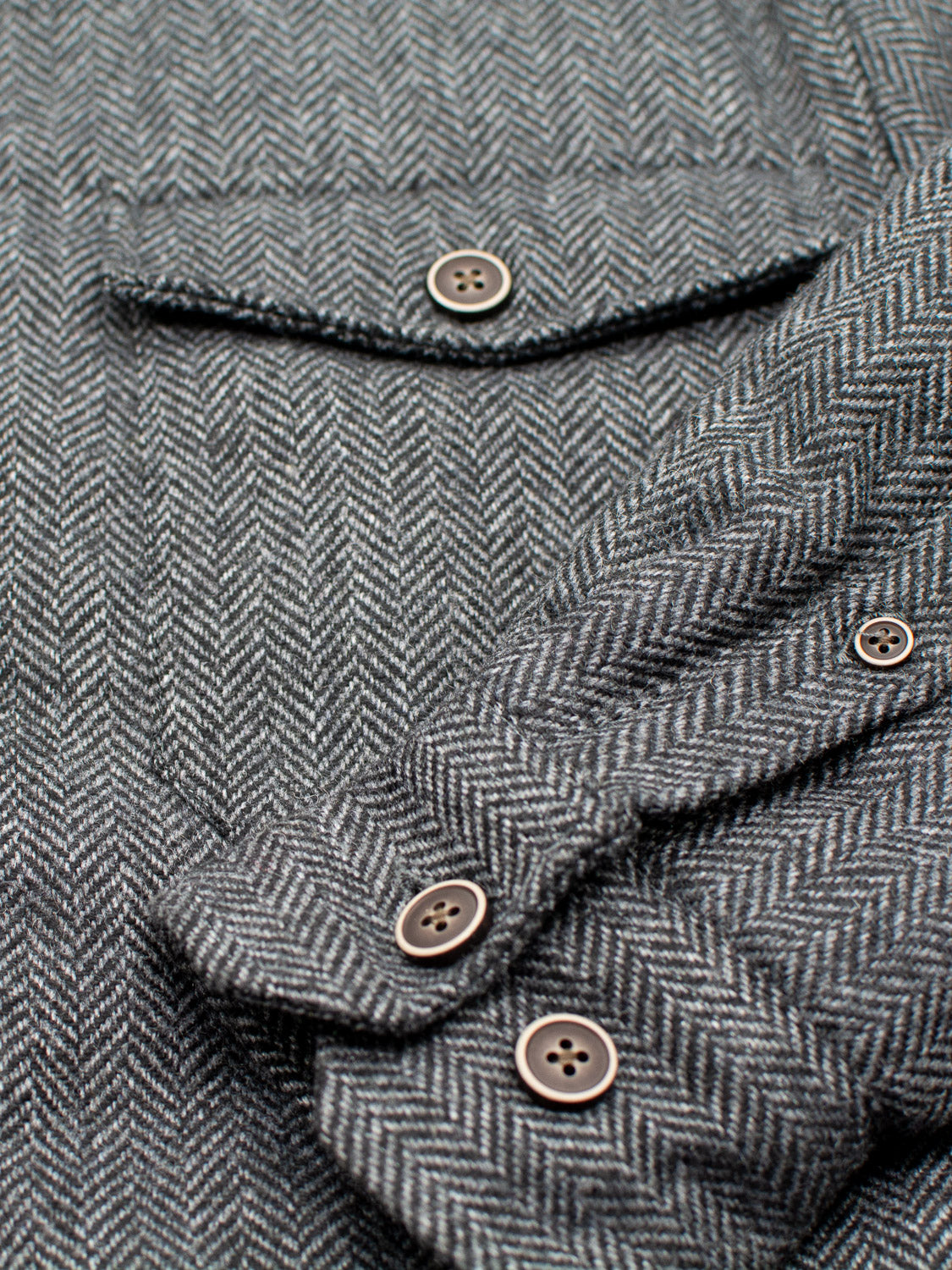 Close-up of the ID Menswear Stuntman Wool Blend Tweed Jacket in gray herringbone, showing a buttoned breast pocket and three-button cuff, with detailed texture and expert stitching for classic outerwear appeal.
