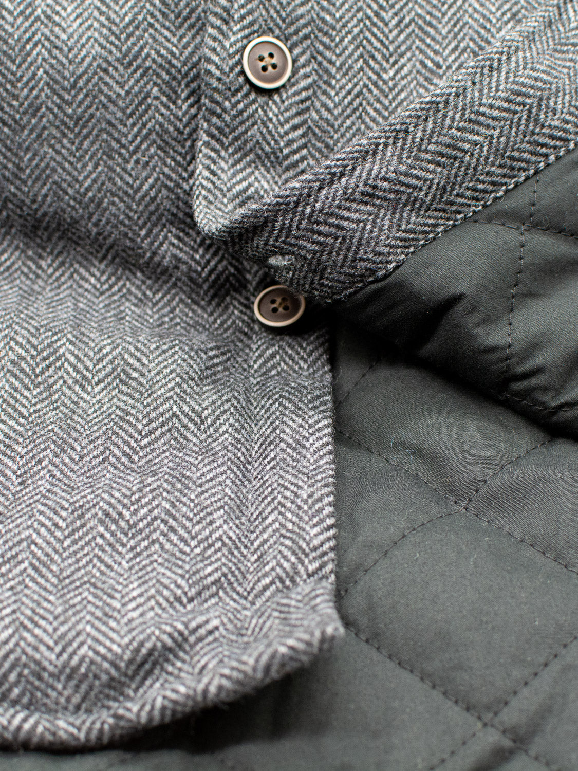 A close-up of the Stuntman Wool Blend Tweed Jacket by ID Menswear shows gray herringbone fabric with buttons beside quilted black lining and collar, highlighting this stylish and timeless outerwear piece.