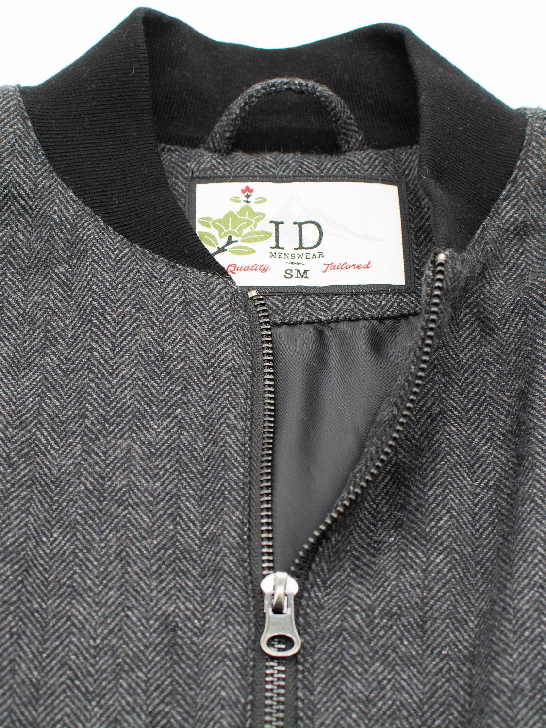 A close-up of the Montauk Wool Blend Jacket in gray herringbone features a visible metal zipper, with the label inside reading ID Menswear and Quality Tailored SM. Made from a wool blend tweed, it offers lightweight comfort with its black collar and textured fabric.