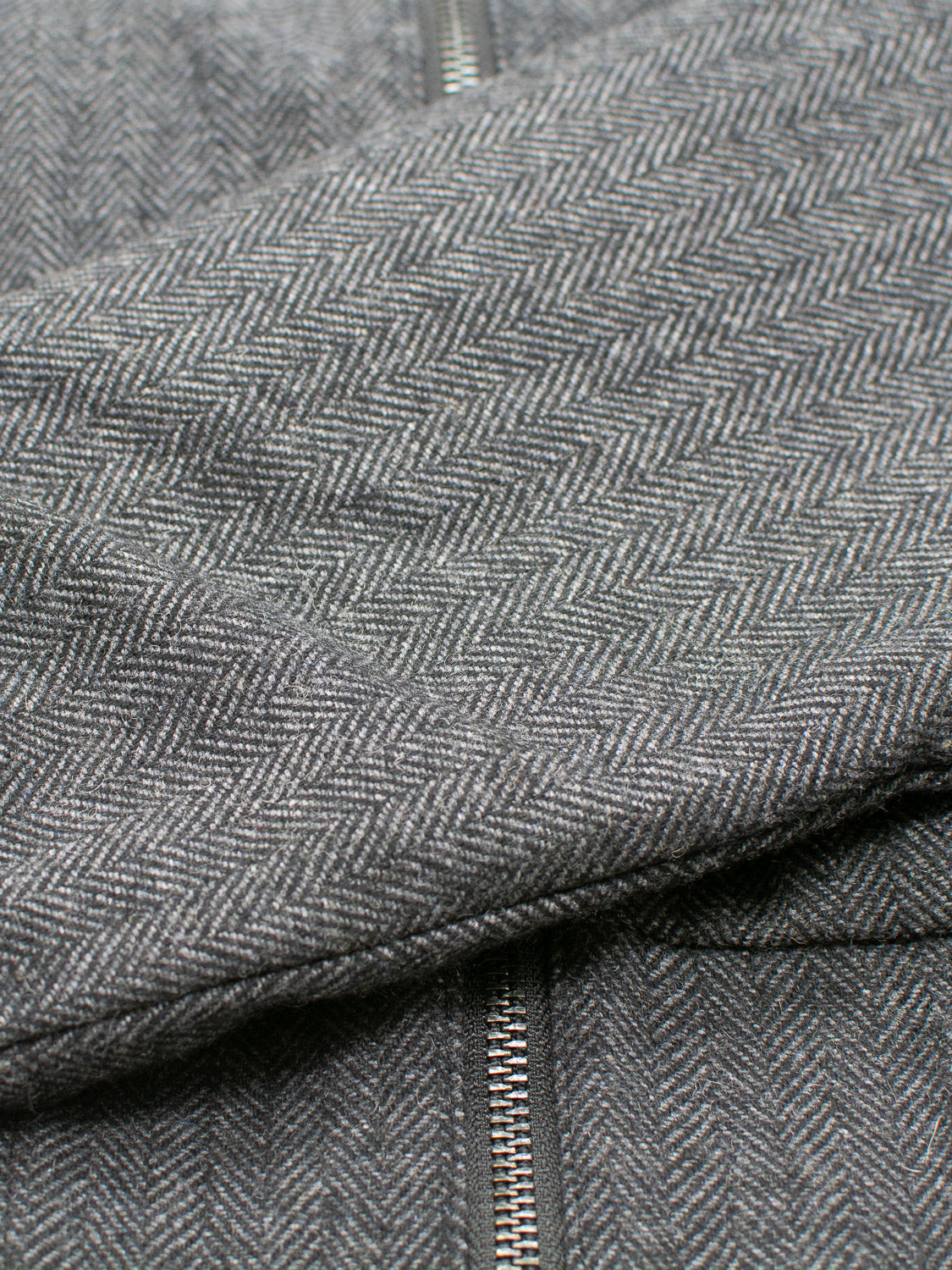 Close-up of a gray herringbone-patterned fabric with a visible vertical metal zipper. The wool blend tweed texture and intricate design evoke the refined charm of the ID Menswear Montauk Wool Blend Jacket.