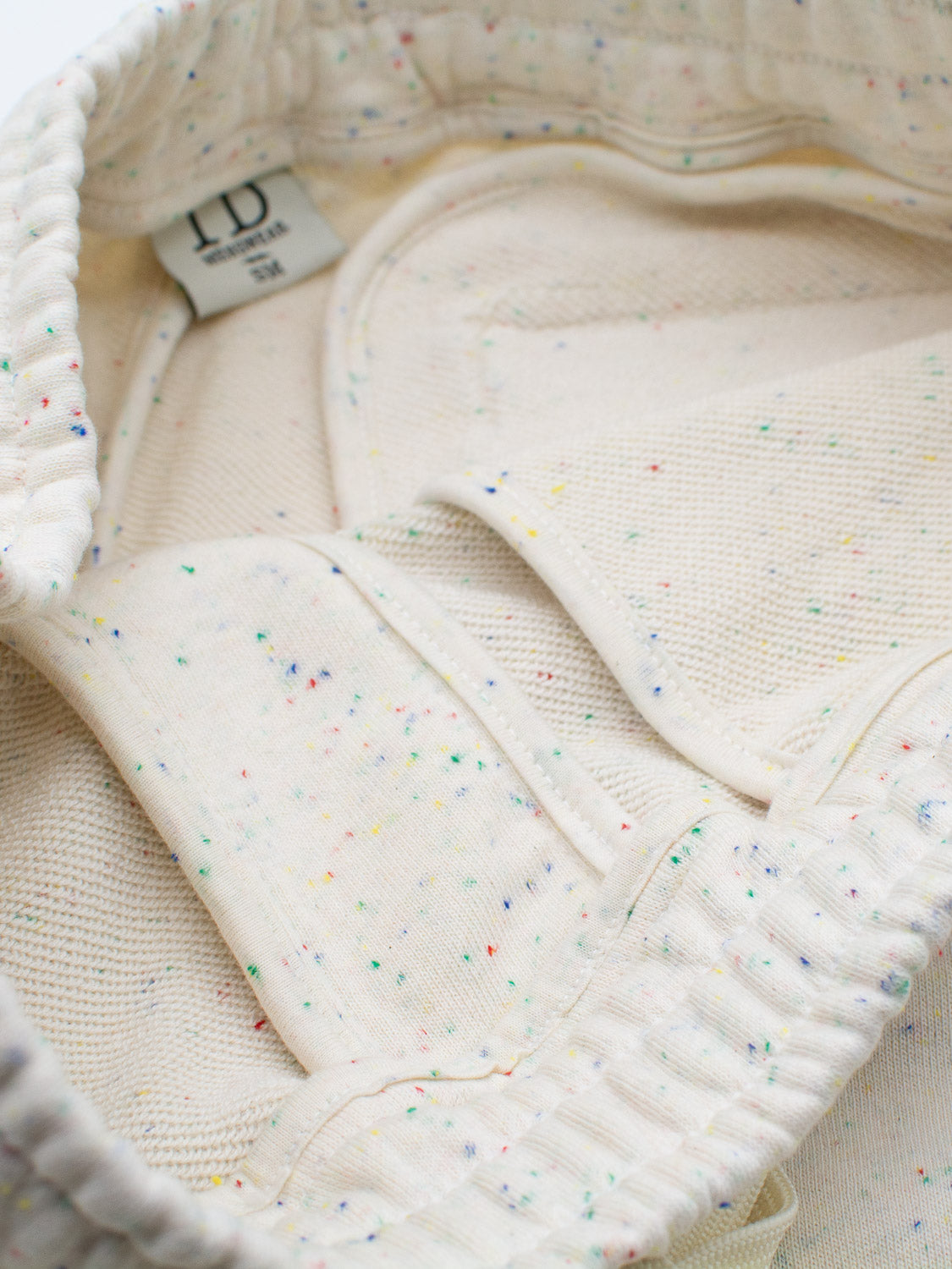 Close-up of Waikiki Speckled Cotton Short by ID Menswear, featuring a cream-colored fabric with colorful specks and elastic edges. A sewn-in tag reads 10 and Made in Italy, showcasing its fashion-forward style, hinting at its use in garments like sweatpants or sweaters.