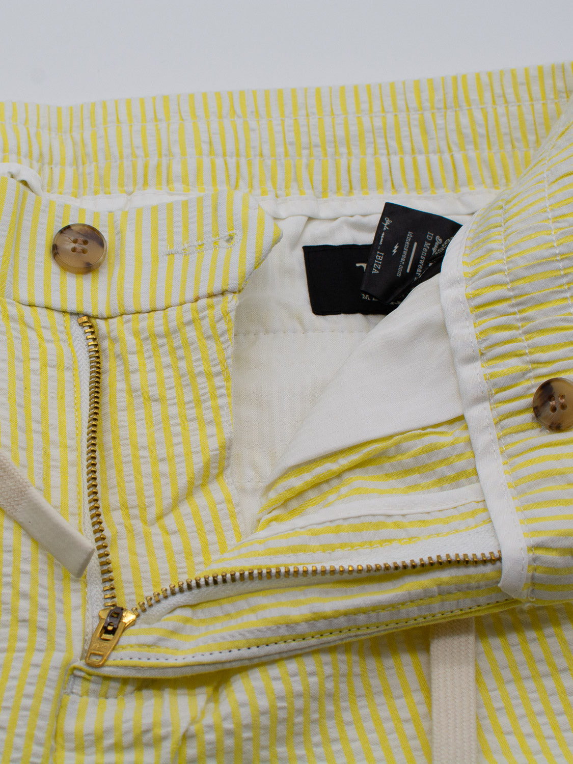 A close-up of ID Menswears Ibiza Seersucker Drawstring Short reveals yellow and white vertical stripes, a partially open zipper, wooden buttons, and visible interior waistband tags—ideal for warm-weather wear.