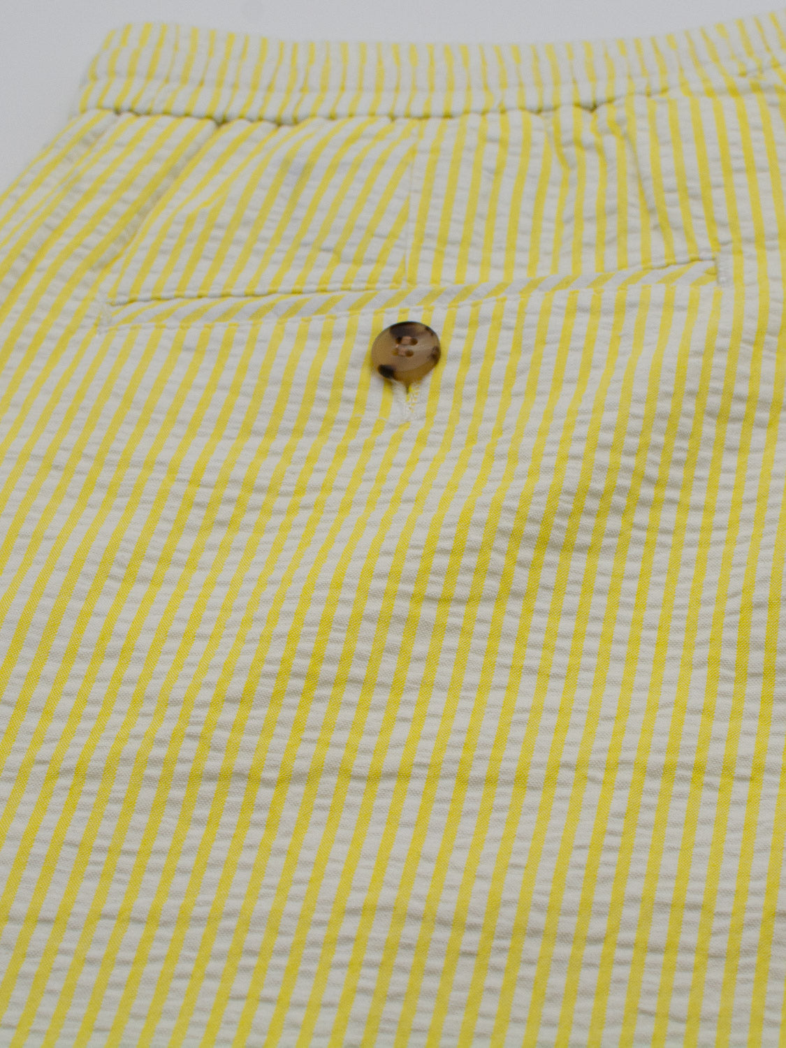 A close-up of yellow and white striped fabric featuring a wooden button and pocket flap suggests the Ibiza Seersucker Drawstring Short by ID Menswear, with vertical stripes offering a summery, casual vibe ideal for warm weather attire.
