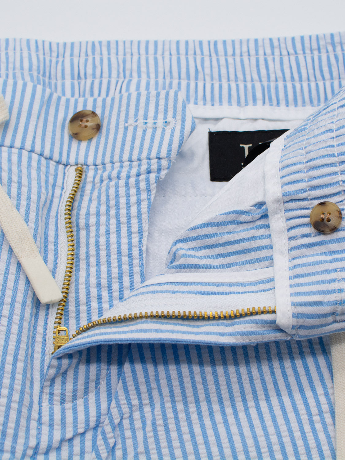 Close-up of ID Menswears Ibiza Seersucker Drawstring Shorts features blue and white stripes with a partially open zipper, buttoned waistband, beige buttons, and a beige drawstring. The lightweight textured fabric is ideal for warm-weather wear.