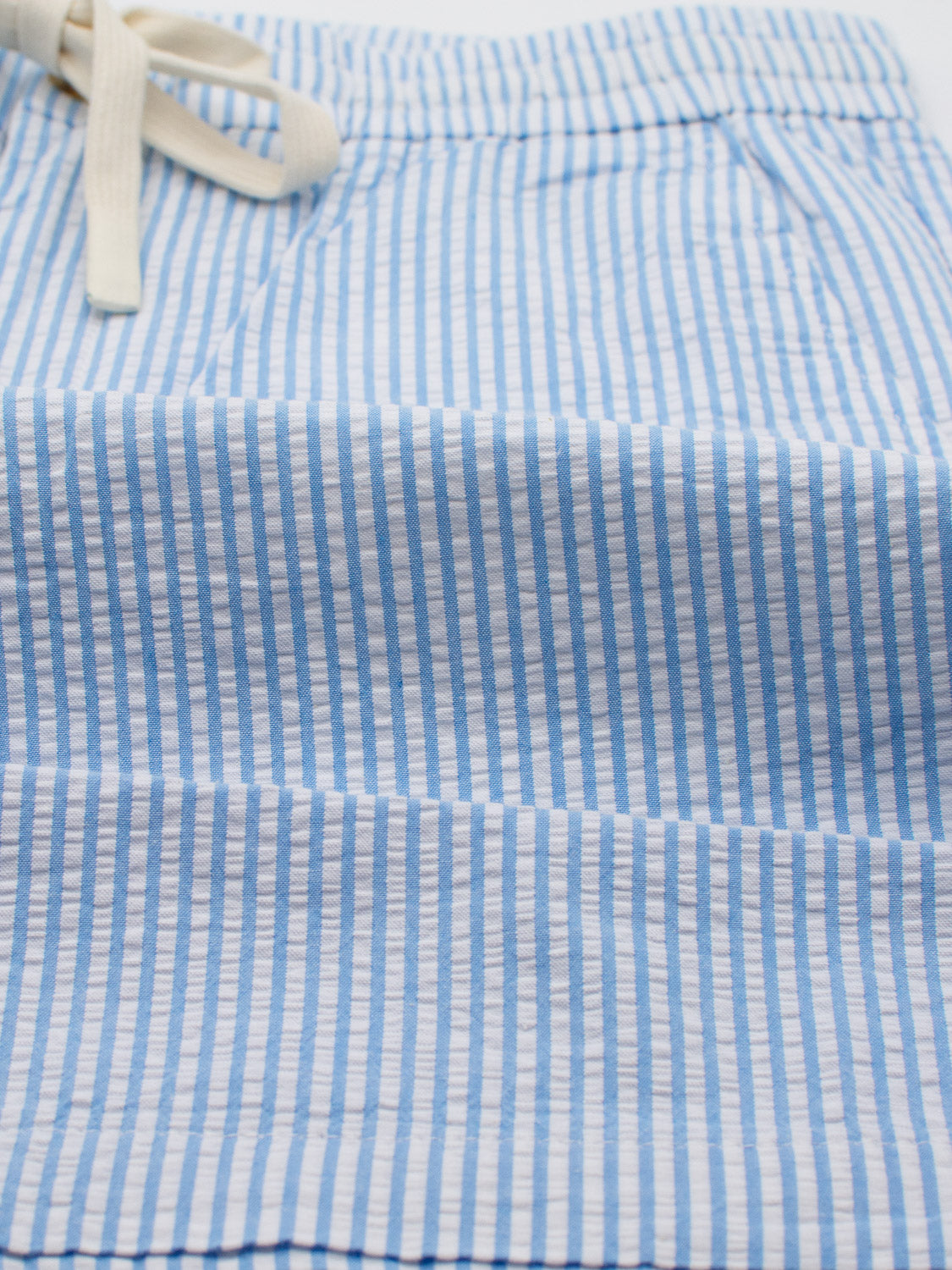 ID Menswears Ibiza Seersucker Drawstring Shorts showcase a close-up of blue and white stripes with an off-white drawstring waist. The airy, puckered texture and vertical stripes make them perfect for warm weather or an Ibiza getaway wardrobe.