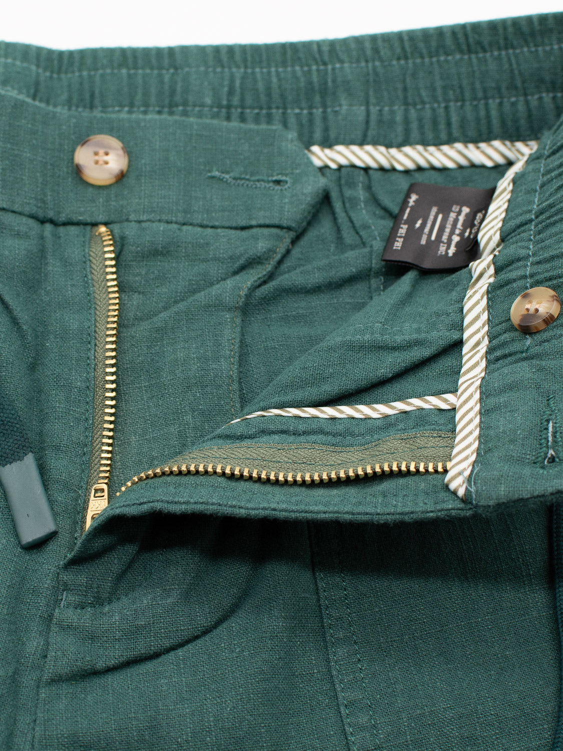 Close-up of green Phi Phi Linen Blend Drawstring Short by ID Menswear, featuring a partially open zipper, tan buttons, and a black label with white text. The herringbone-textured fabric has an elastic waistband for comfort.