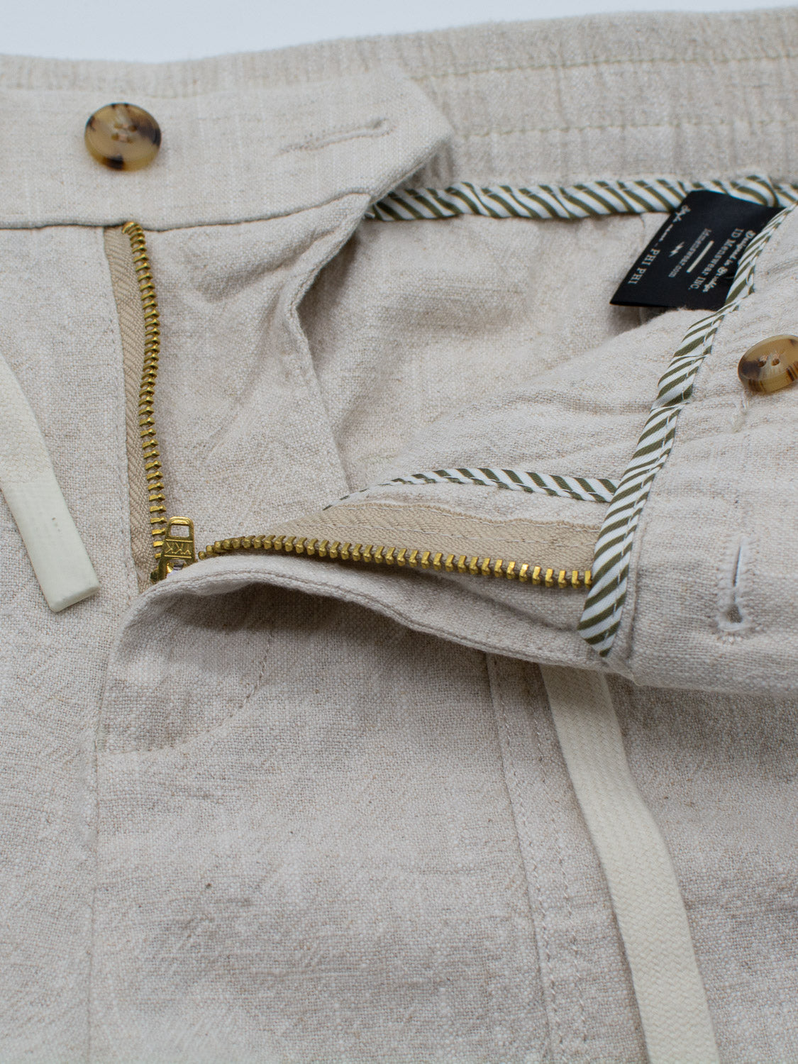 Close-up of ID Menswears Phi Phi Linen Blend Drawstring Short featuring a 6 inseam, beige fabric, metal zipper, light drawstring, and button. The elastic waistband has a striped lining detail and visible clothing care tag.