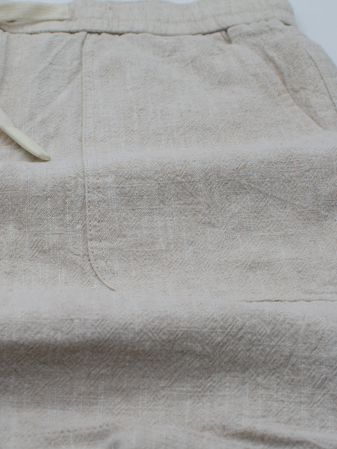 A close-up of ID Menswears Phi Phi Linen Blend Drawstring Shorts showcases their beige, subtly textured pattern with an elastic waistband and drawstring. The 6 inseam enhances comfort, and the slightly wrinkled look highlights the natural fiber composition.